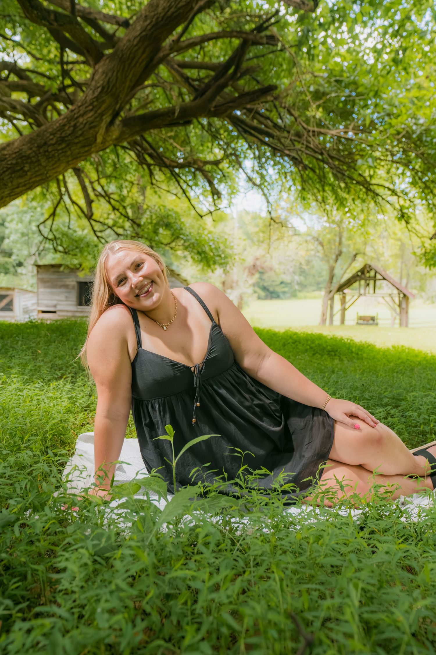 Smiling blonde woman in black dress sitting on grass blanket under large tree in lush green park.