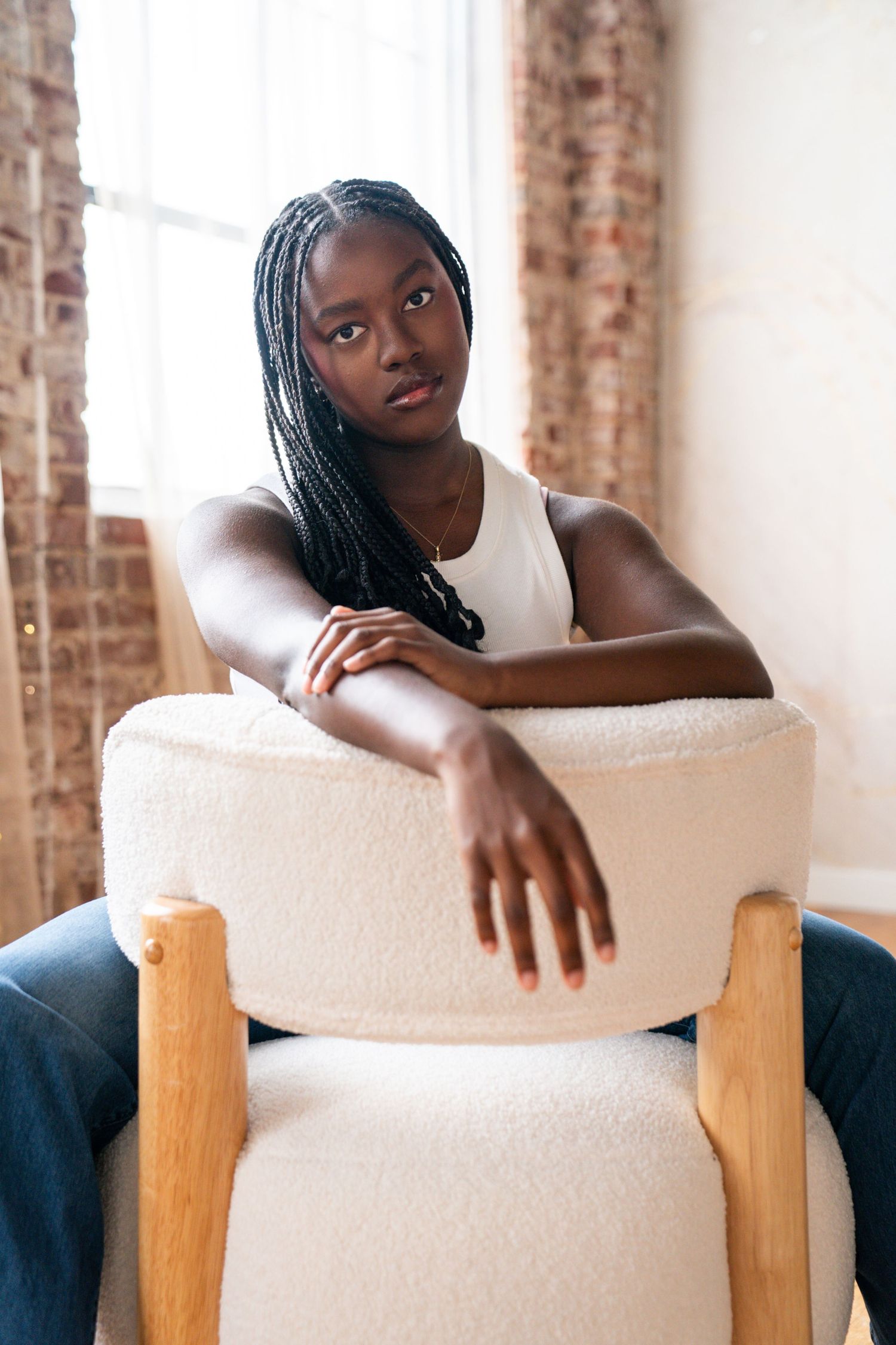 Young Black woman with long braids sits in a white boucle armchair, wearing a white tank top, near a brick wall window.