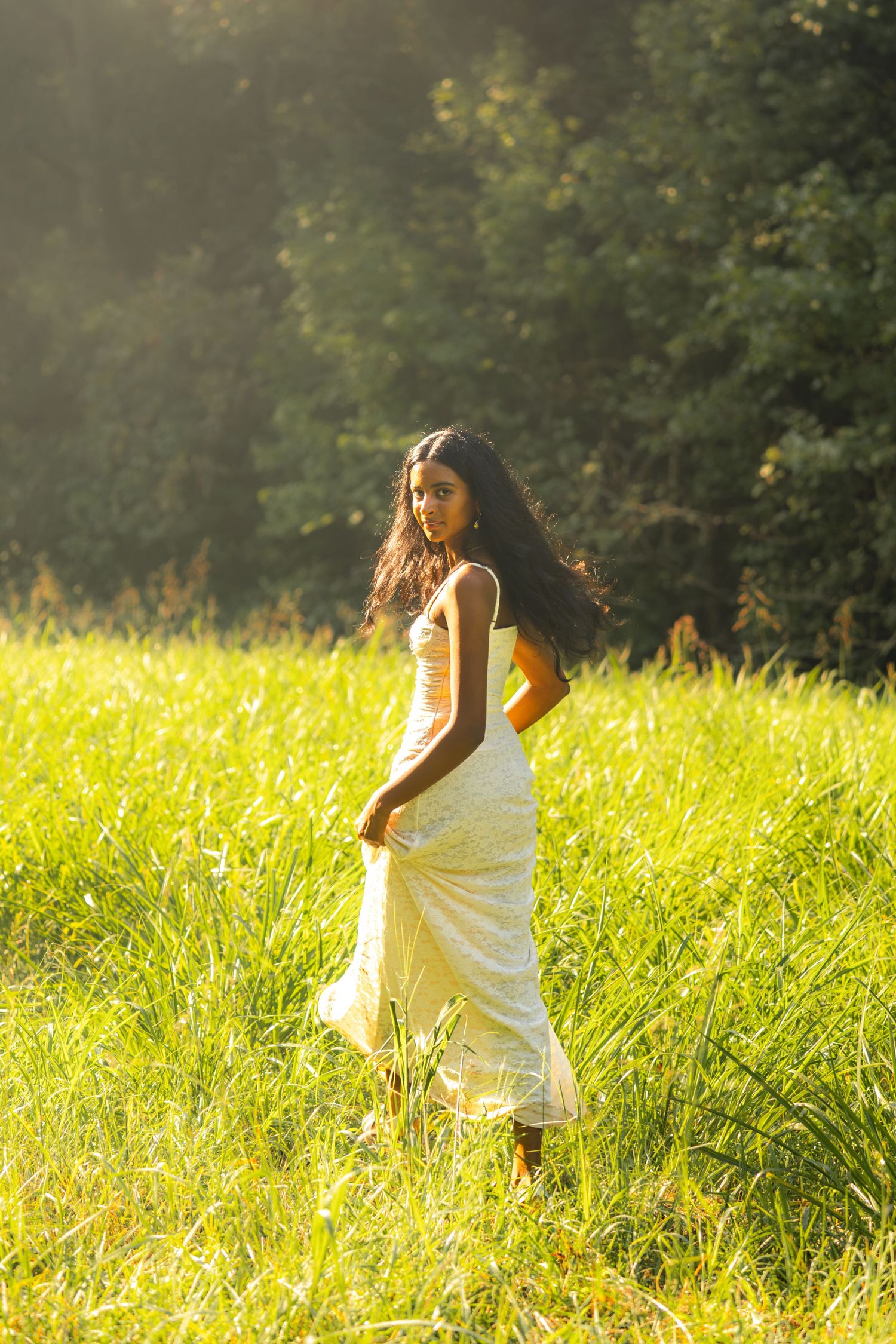Young woman in white dress standing in sunlit green meadow with trees, golden hour photography.