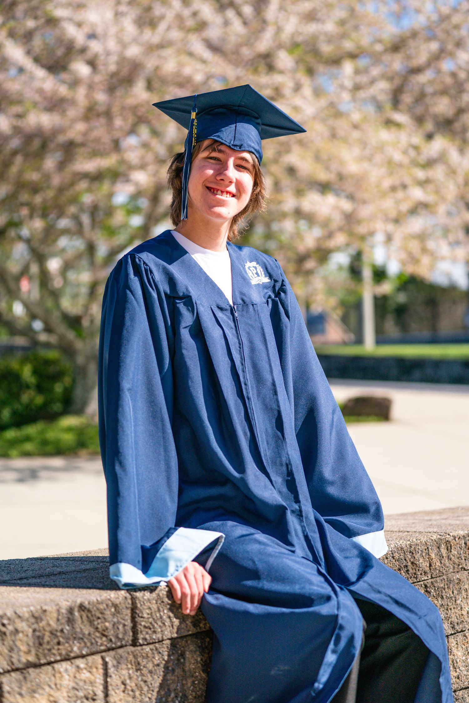 A smiling graduate wearing a blue cap and gown sits on a stone wall in front of blooming trees.