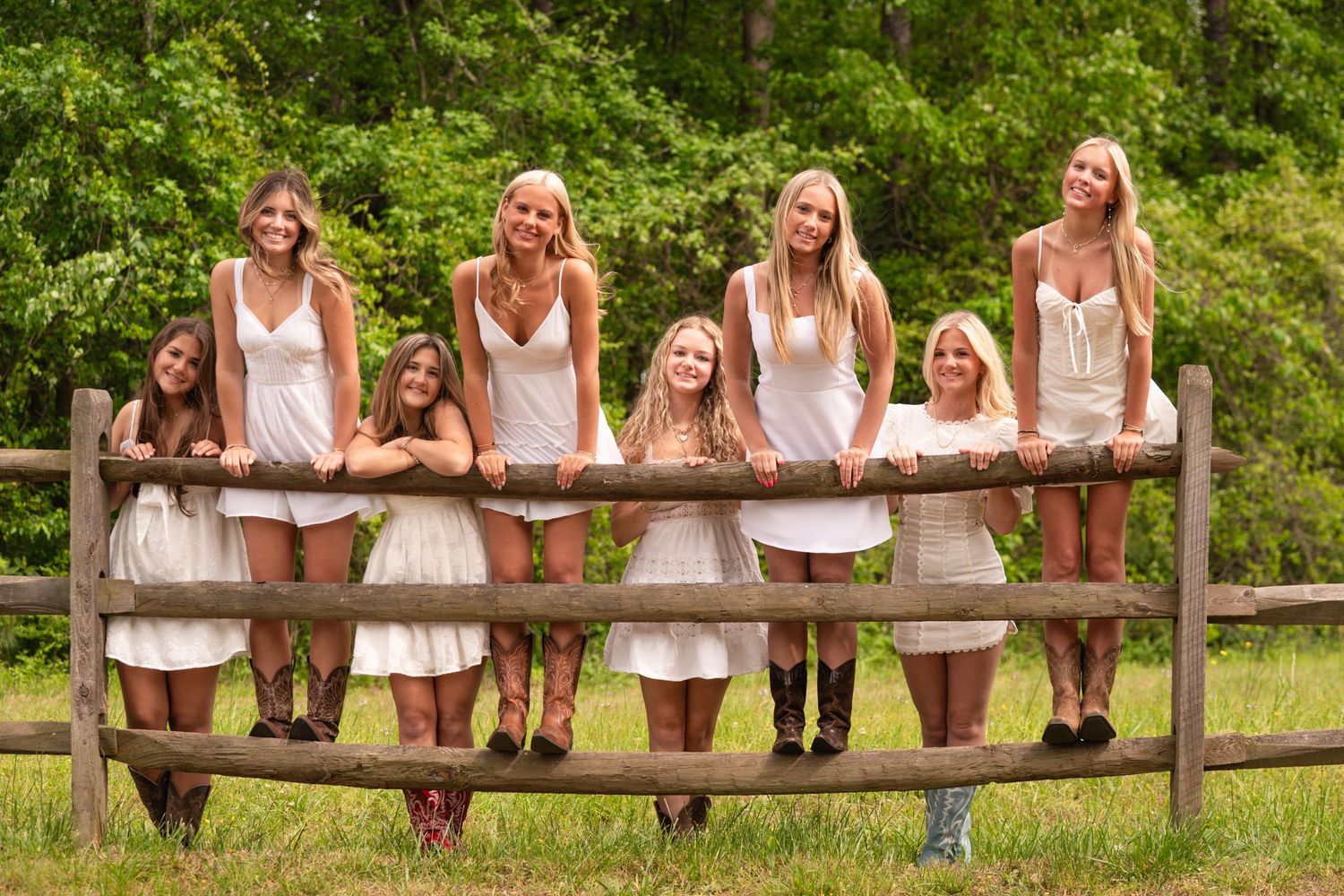 Group of young women in white dresses posing together along a rustic wooden fence in a lush green outdoor setting.