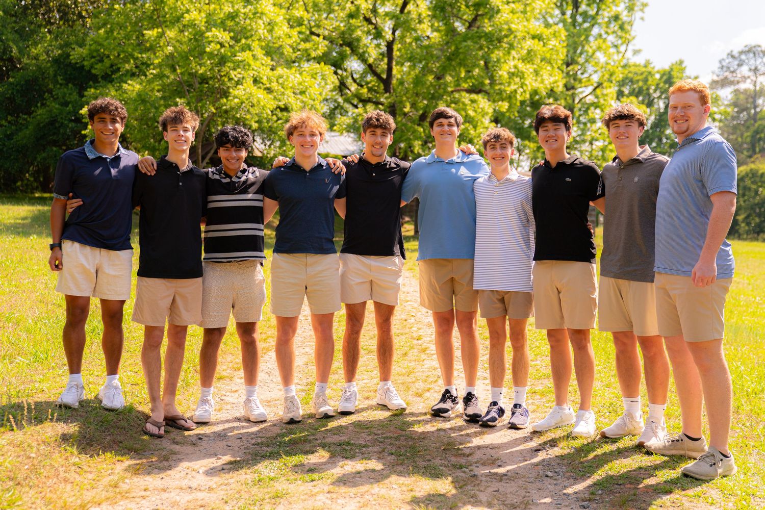 Group of nine young men posing together outdoors in khaki shorts on a sunny day in a park setting.