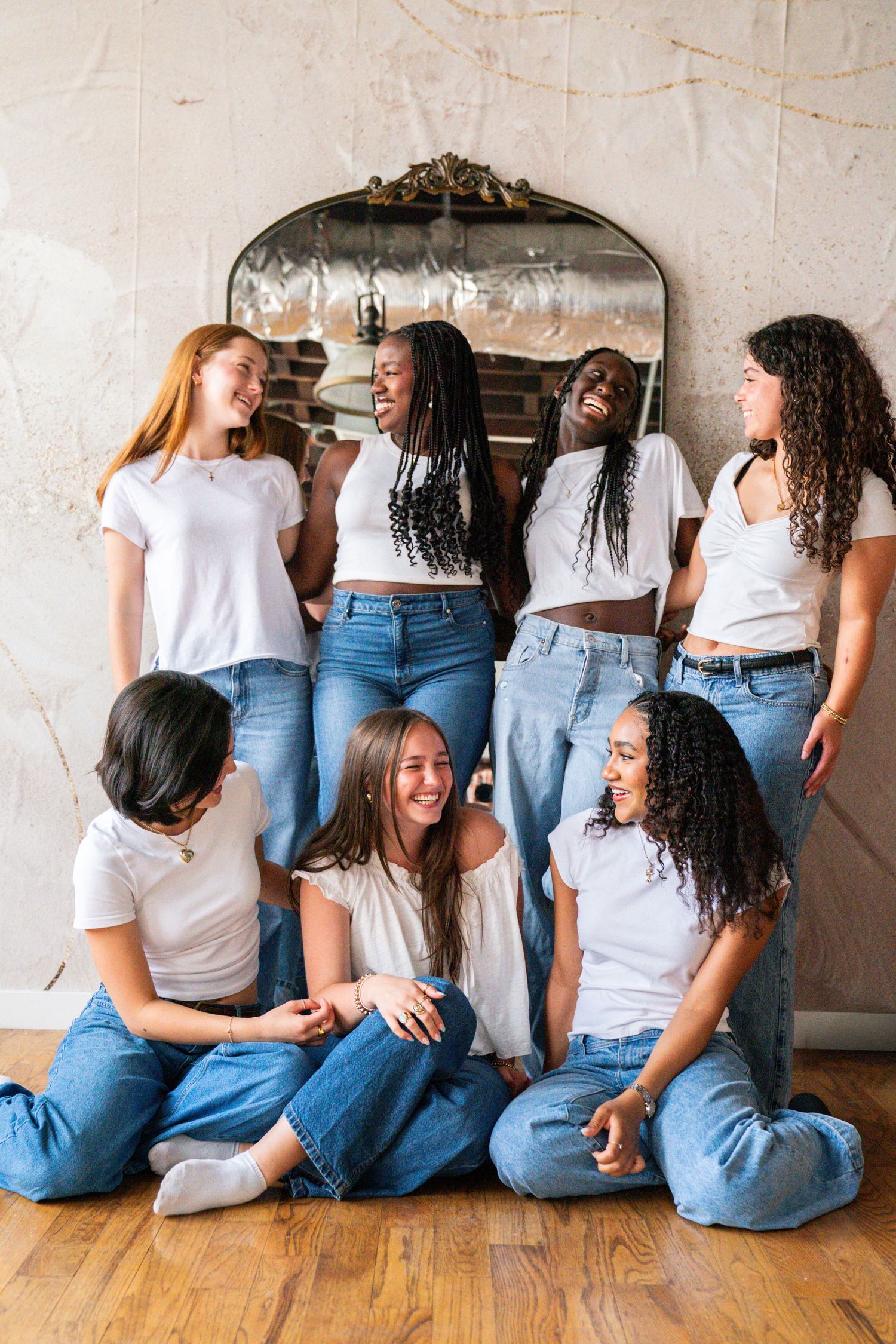 Diverse group of seven women in white tops and jeans laughing together in front of an ornate mirror.