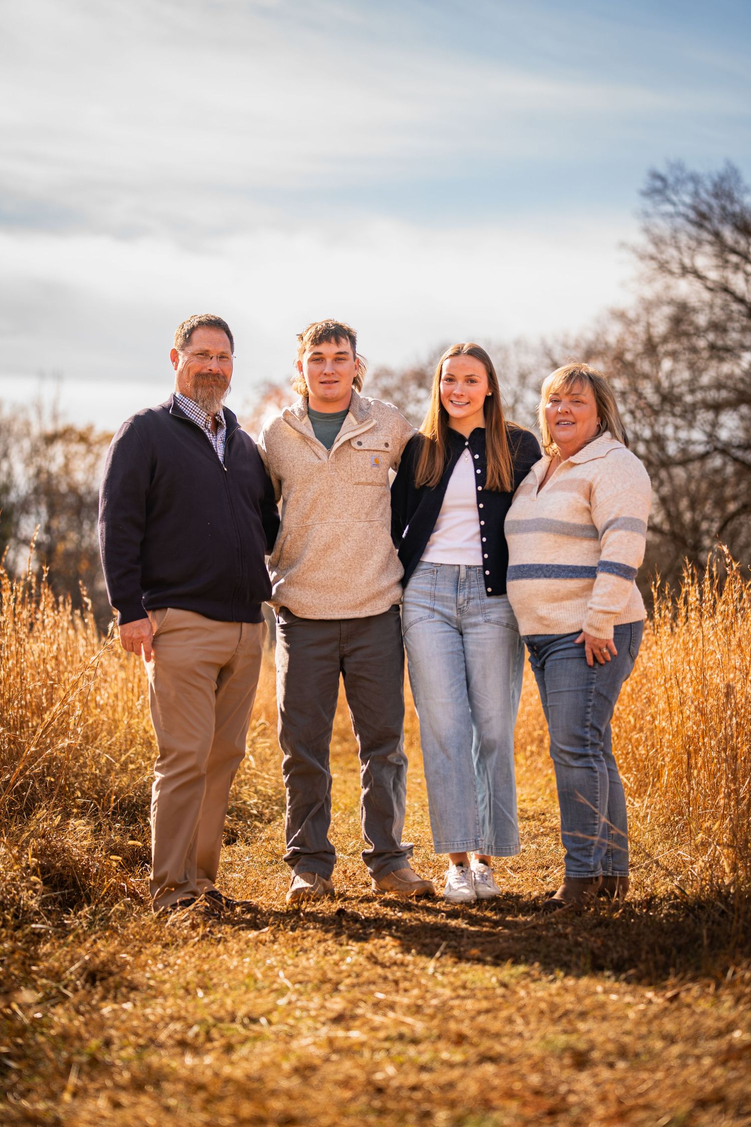 Family of four smiling outdoors in a golden field during autumn, posing together under a partly cloudy sky.