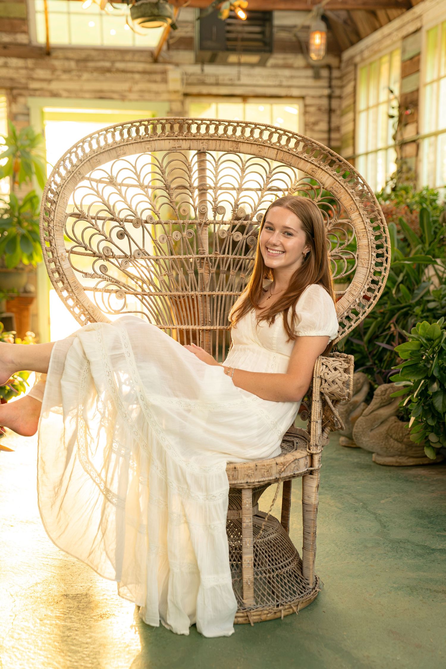 Young woman in white dress sitting in wicker peacock chair, smiling in sunlit greenhouse with lush green plants.