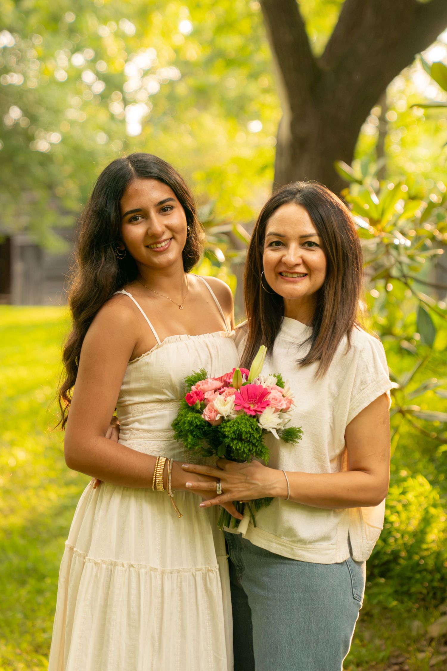 Two women smiling outdoors holding a pink flower bouquet, standing together in a lush green garden setting.