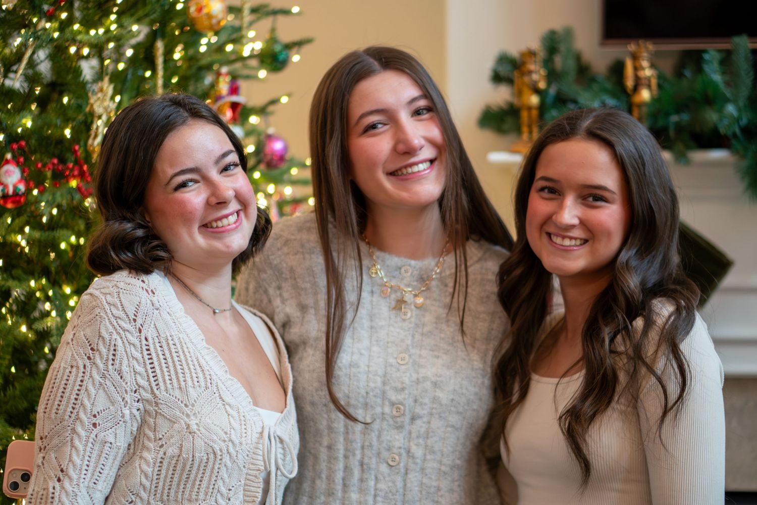 Three friends in cozy sweaters smile together in front of a decorated Christmas tree during the holiday season.