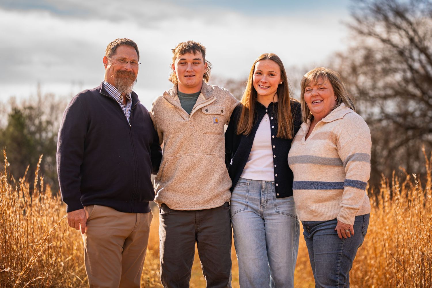 Family group poses together outdoors in front of golden wheat field during autumn sunset.
