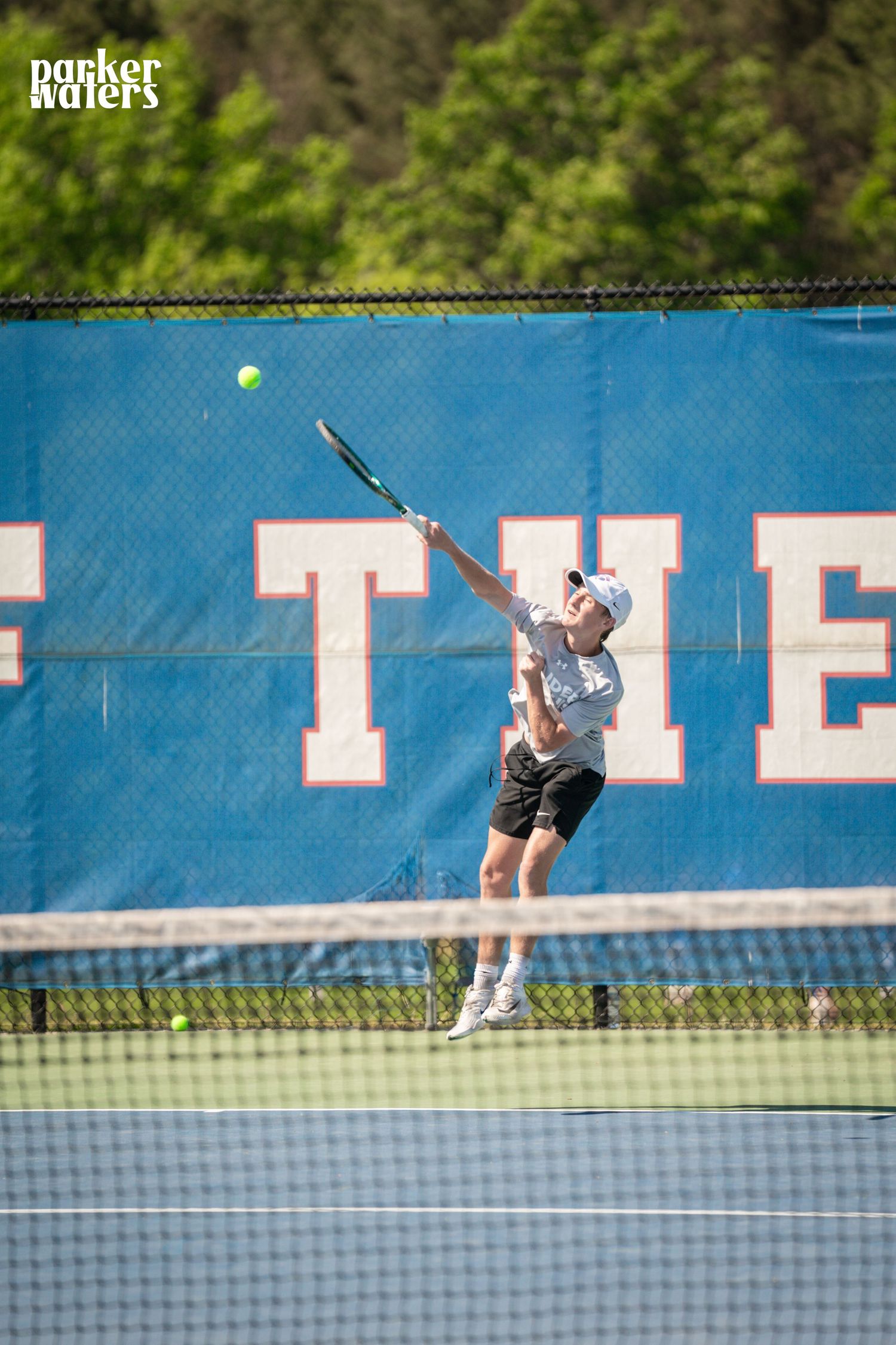 A tennis player leaps to serve on an outdoor court with a blue banner backdrop.