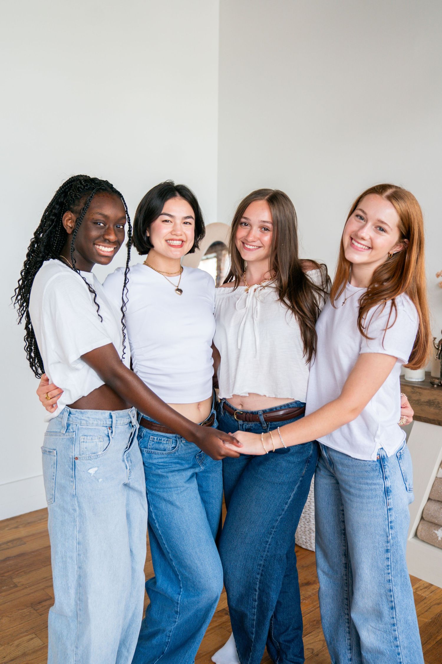 Four diverse young women in white tops and jeans smiling and embracing together indoors.