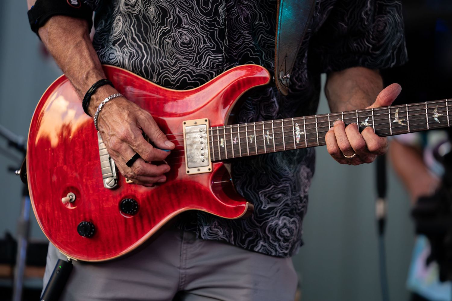 Musician playing a red PRS electric guitar on stage, wearing a patterned shirt and bracelet.