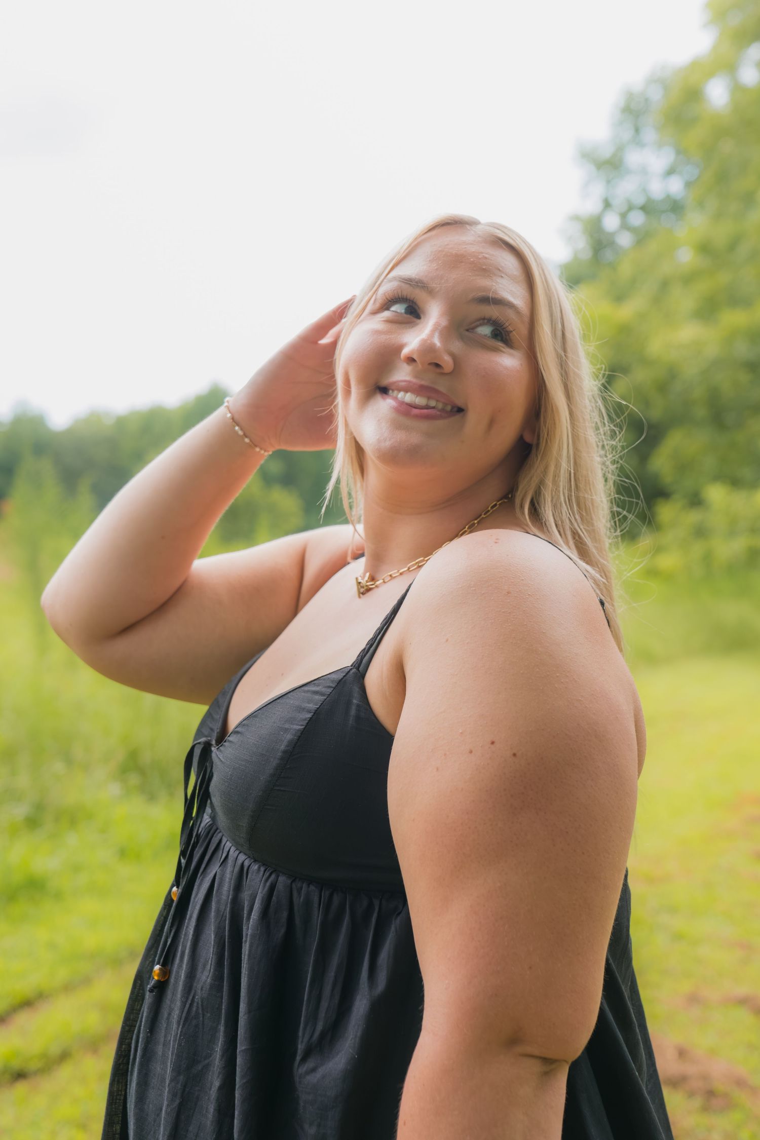 Smiling blonde woman in black sundress poses outdoors in green nature setting, hand raised near face.