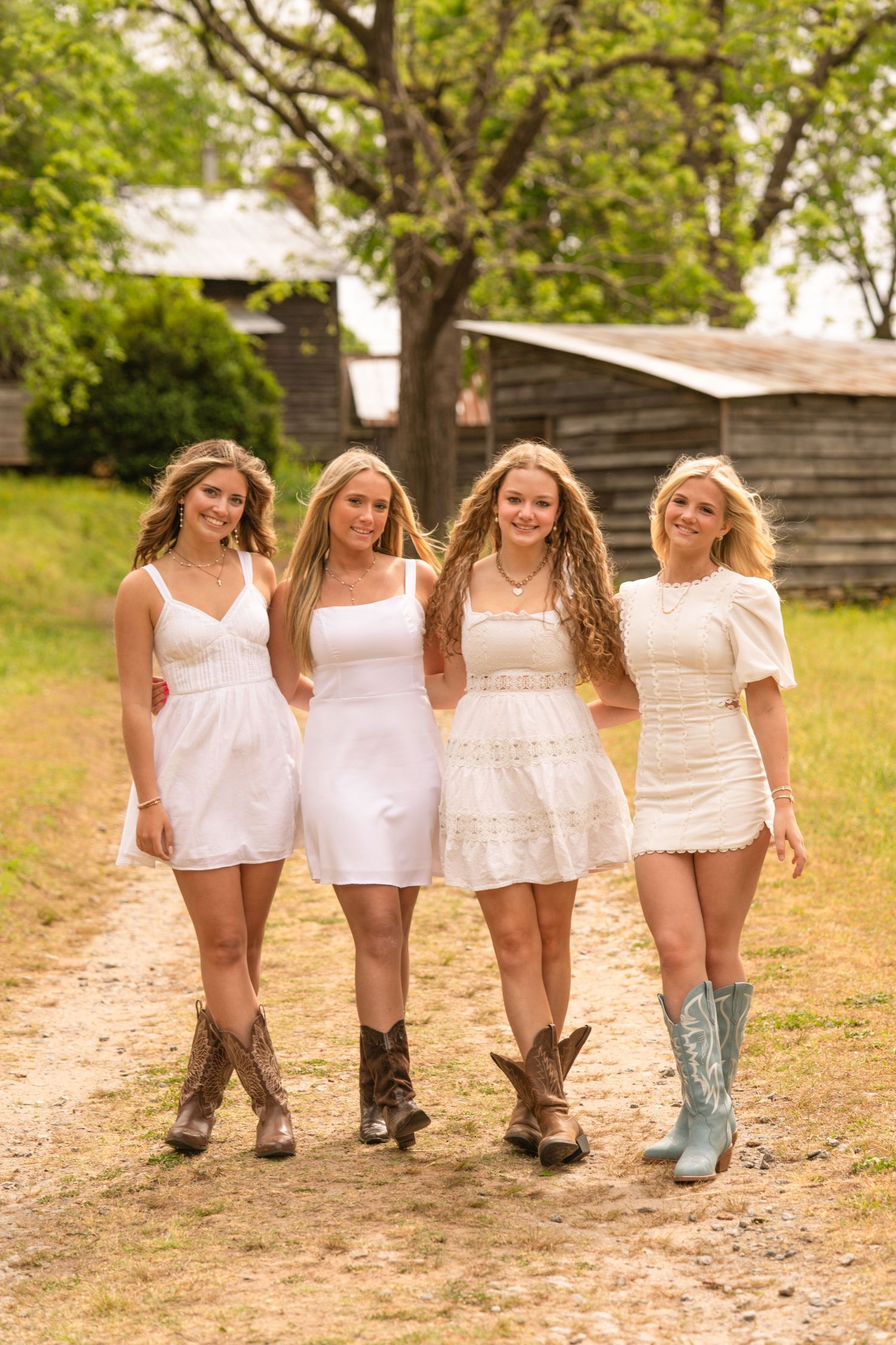 Four young women in white dresses and cowboy boots walking together on a dirt path near a rustic barn.