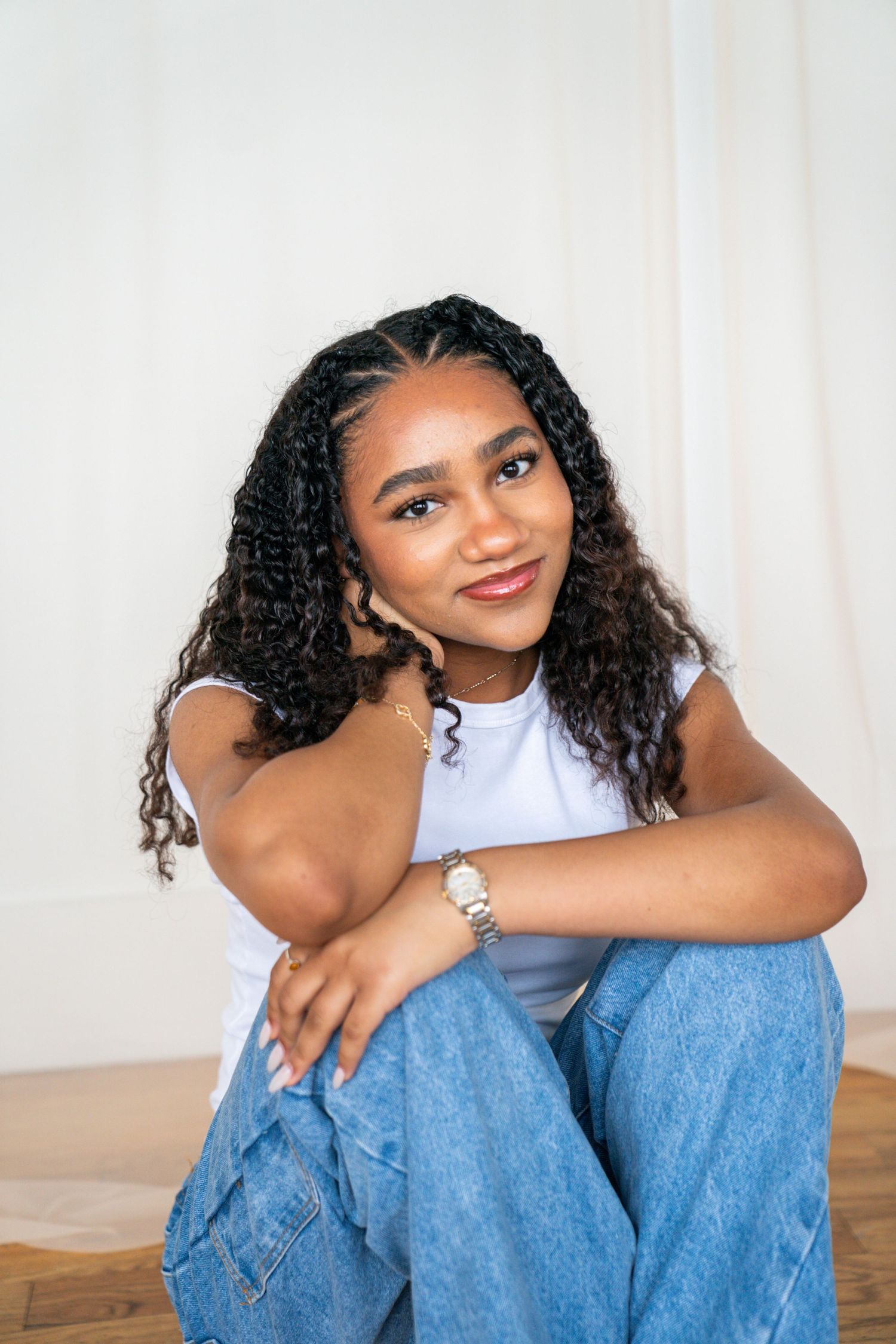 Young Black woman with curly hair sits on floor in white tee and jeans, smiling confidently at camera.