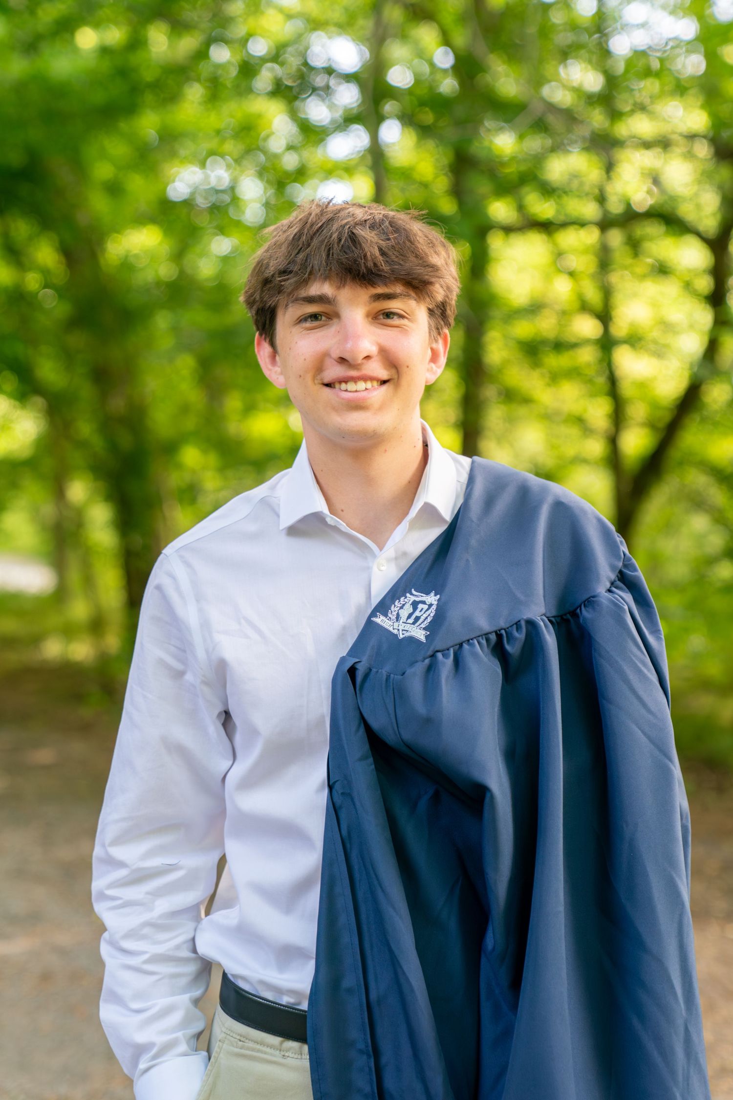 Smiling young man in white shirt holding navy blue graduation gown outdoors among green trees.