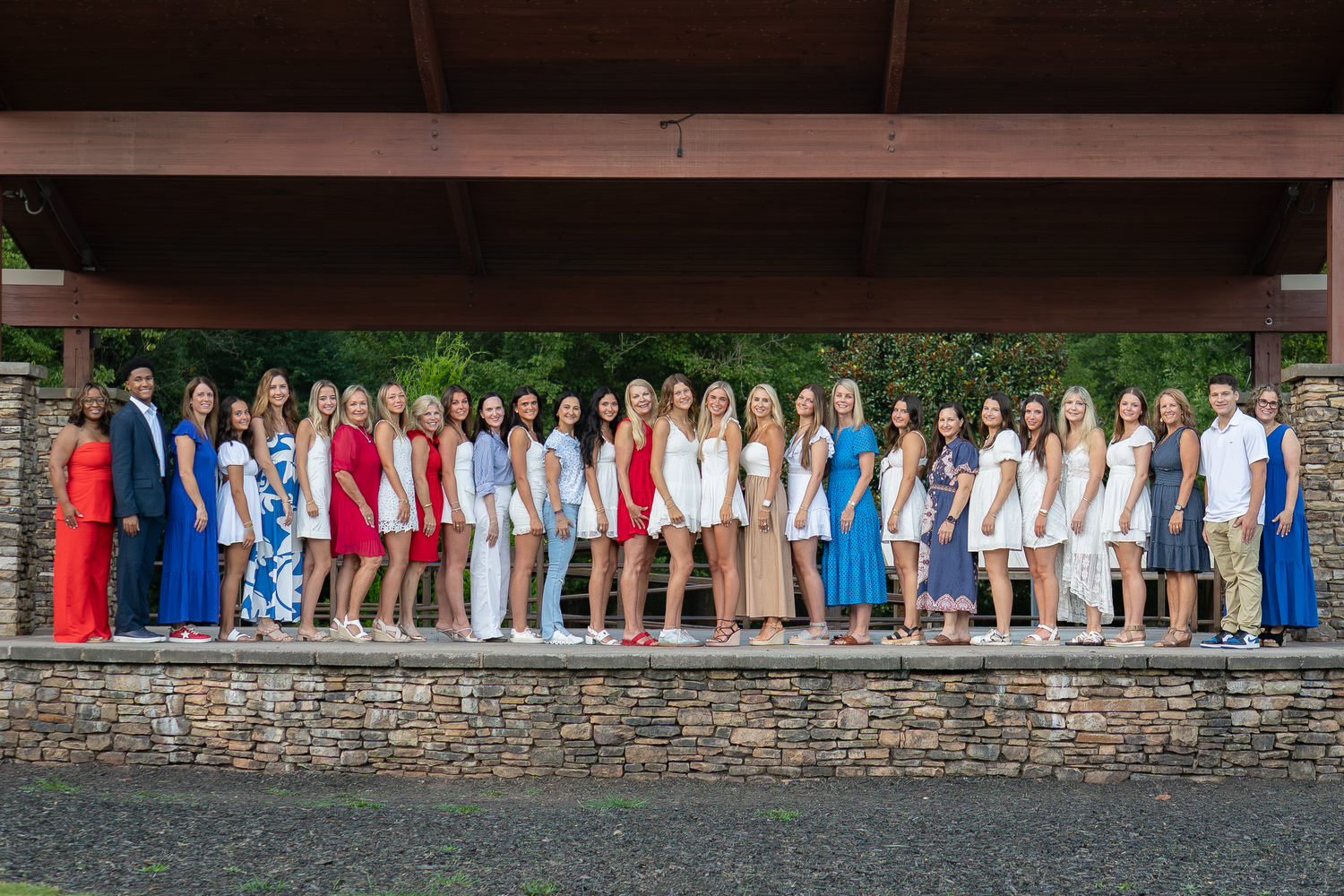 Line of students in colorful outfits posed against stone wall for group photo.