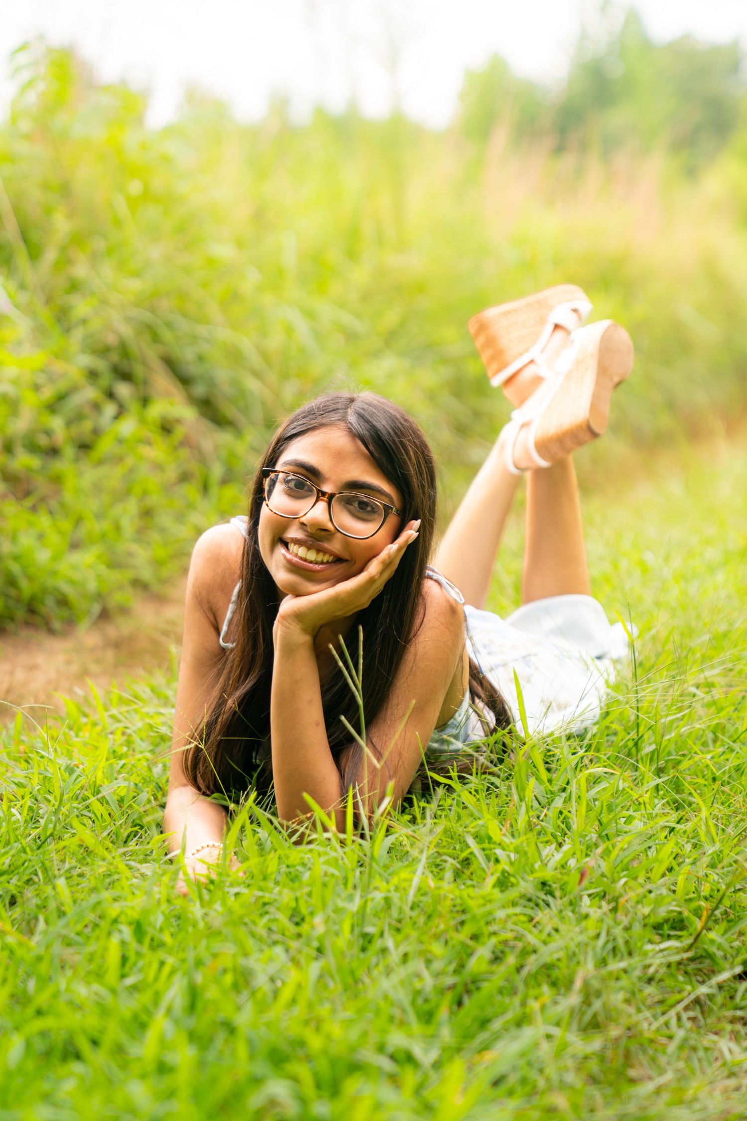 Young girl with glasses lying on green grass in a park, smiling cheerfully at the camera on a sunny day.