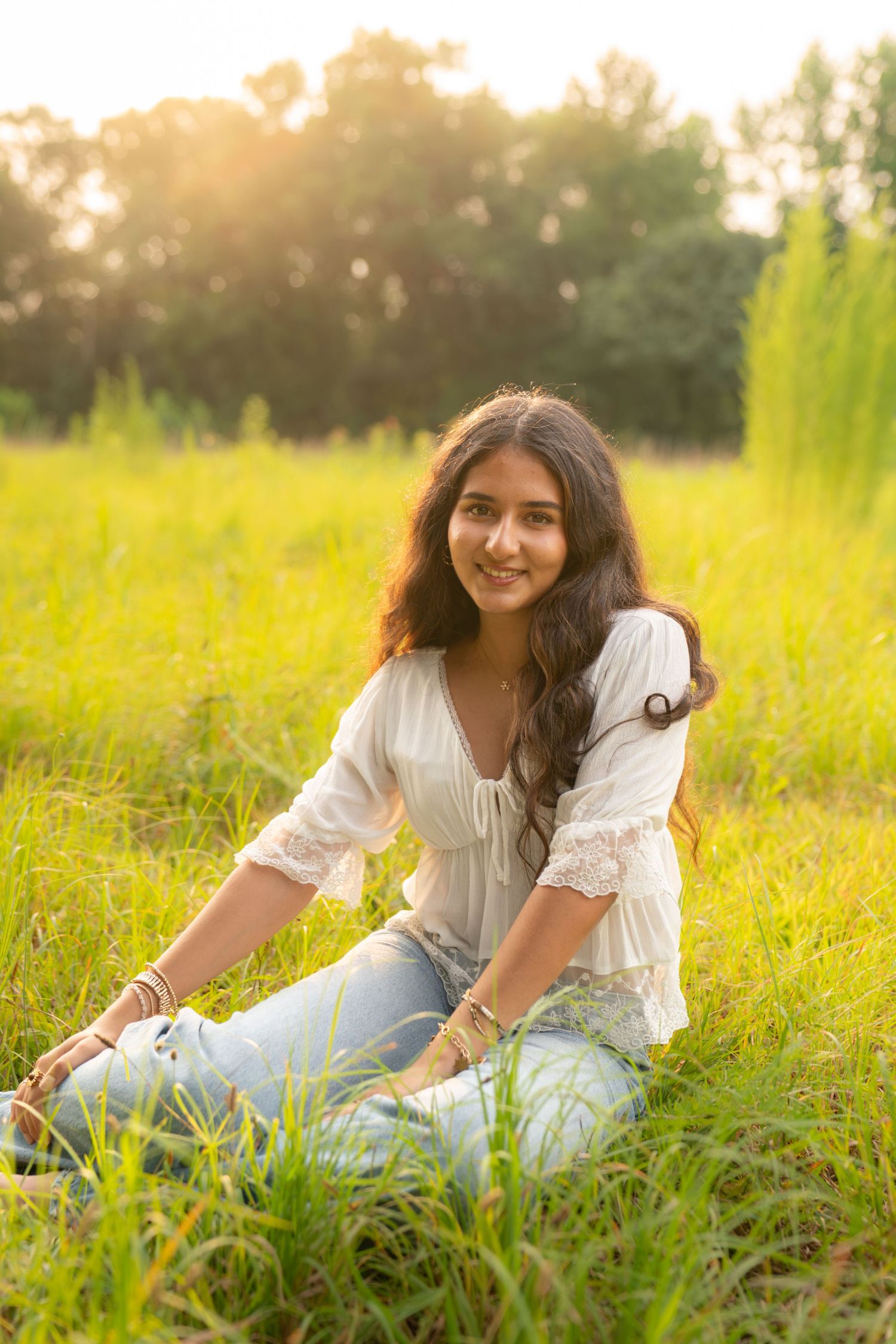 Young woman sitting in lush green grass field at golden hour, wearing white lace top and jeans, smiling warmly.