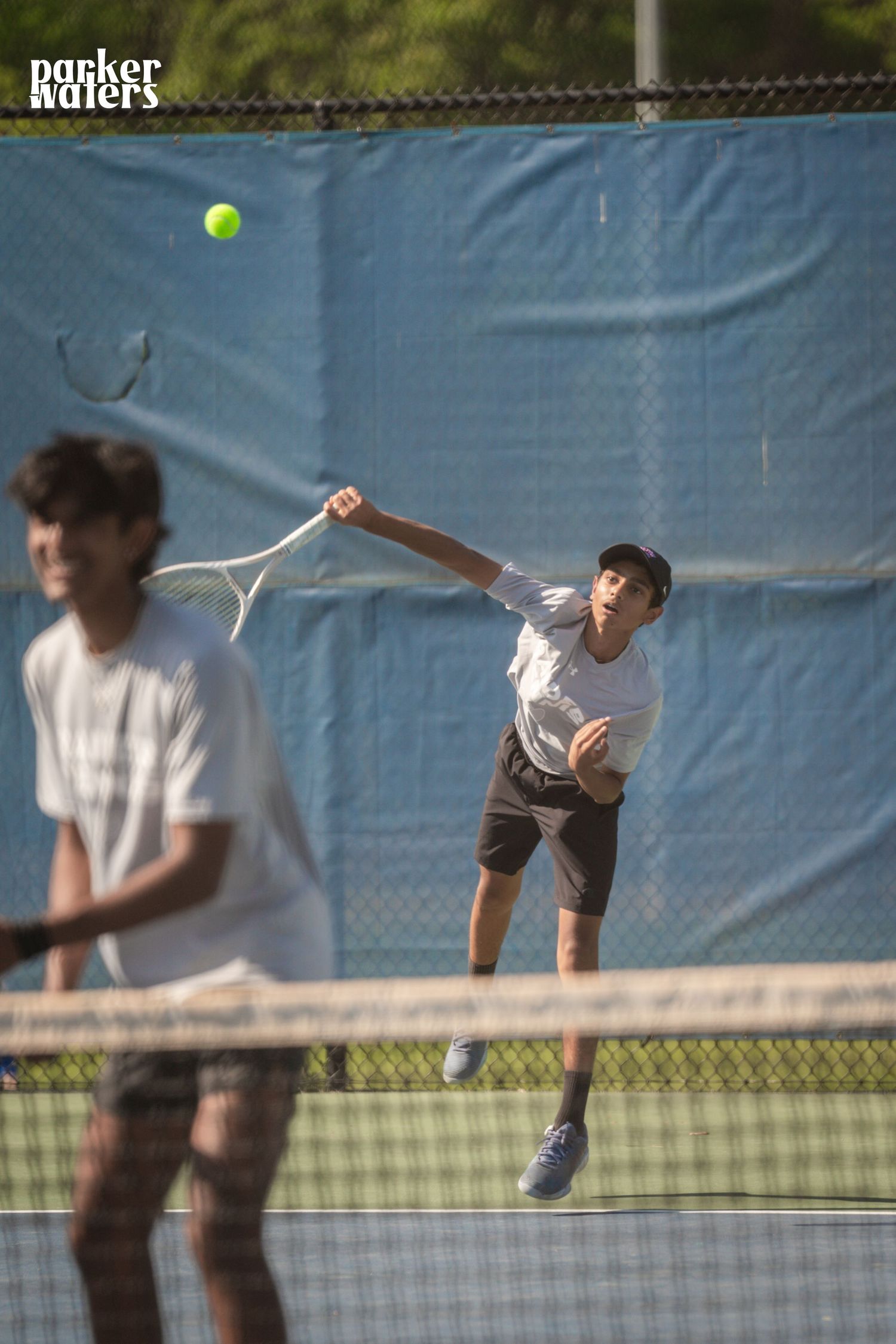 Young tennis player serving ball mid-match on outdoor court, doubles partner visible in foreground.