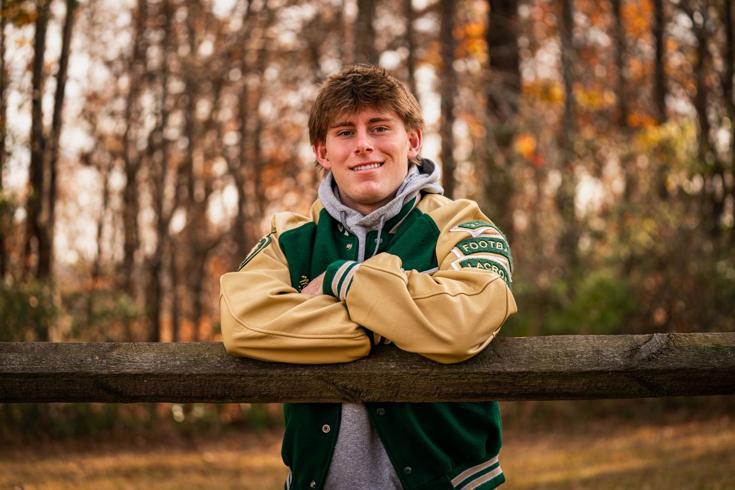 A student in a green and gold varsity jacket leans on a wooden railing during autumn with fall foliage in the background.