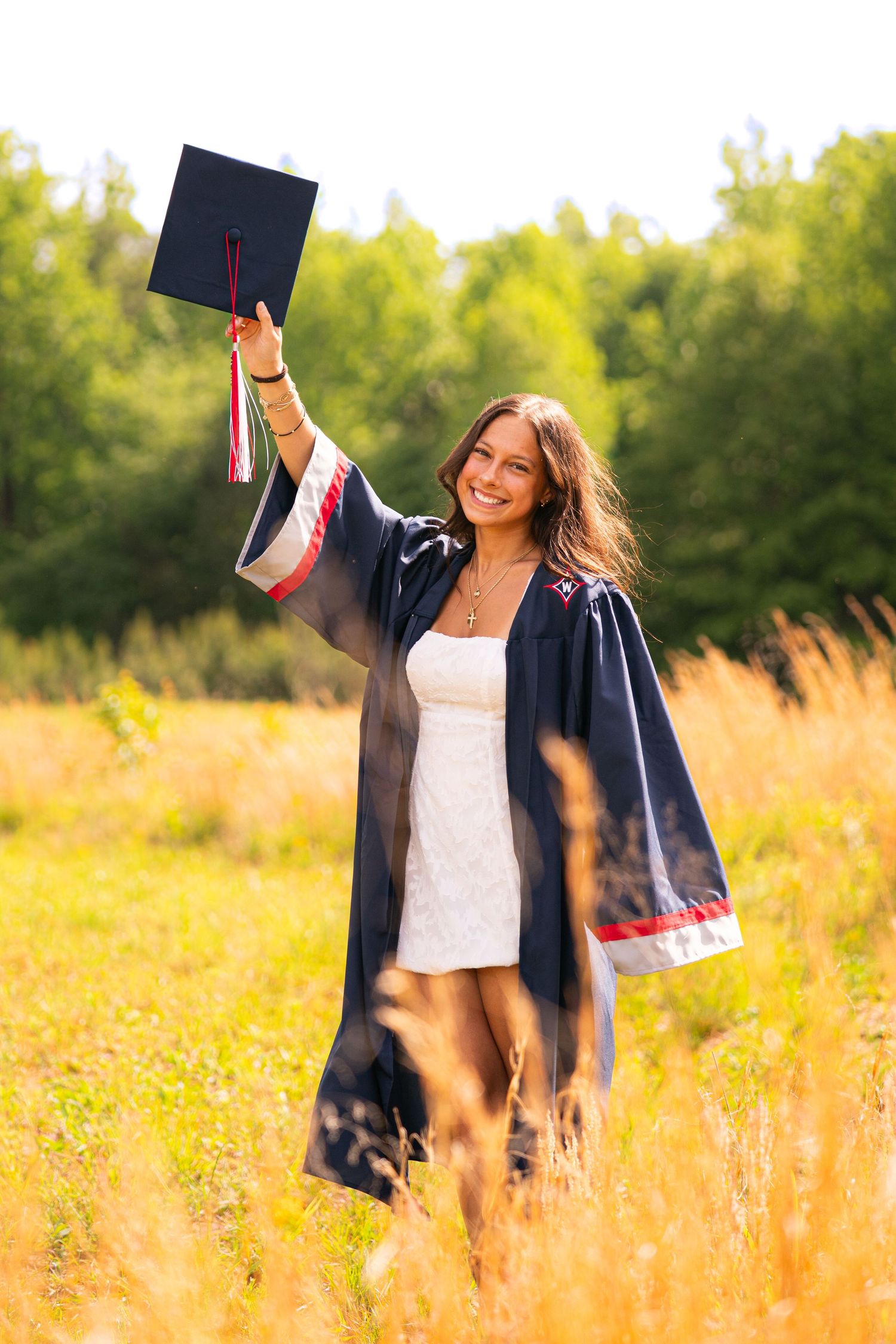 Smiling graduate in cap and gown raising mortarboard in sunny field, celebrating graduation outdoors.