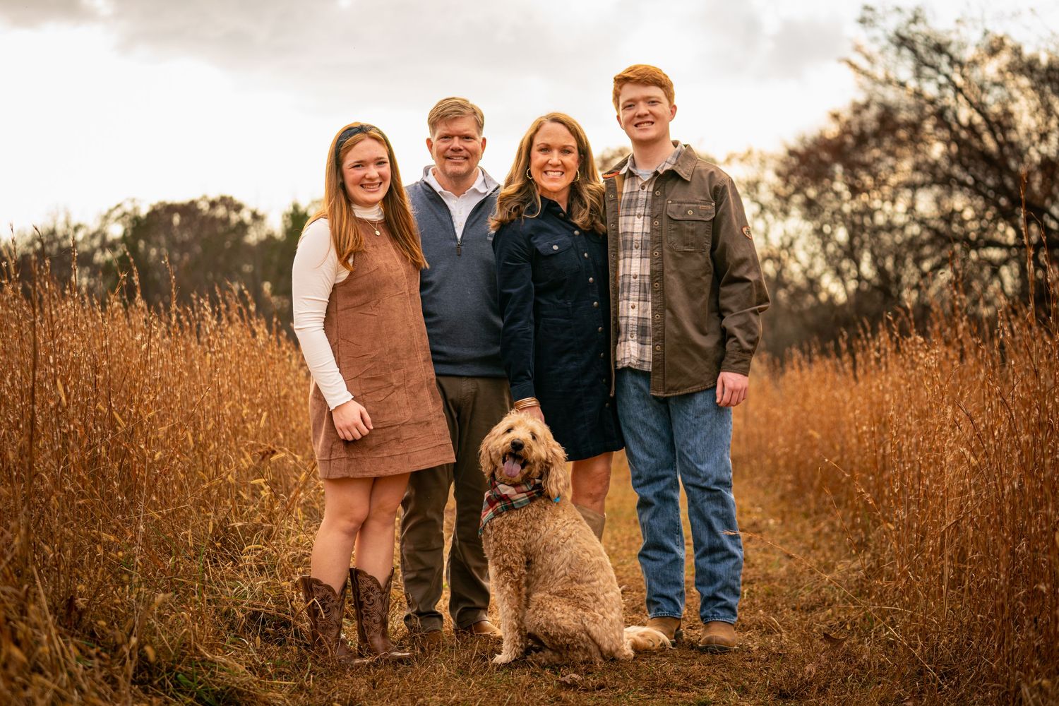 Family of four smiling outdoors in a golden field during autumn, with a fluffy dog sitting at their feet.