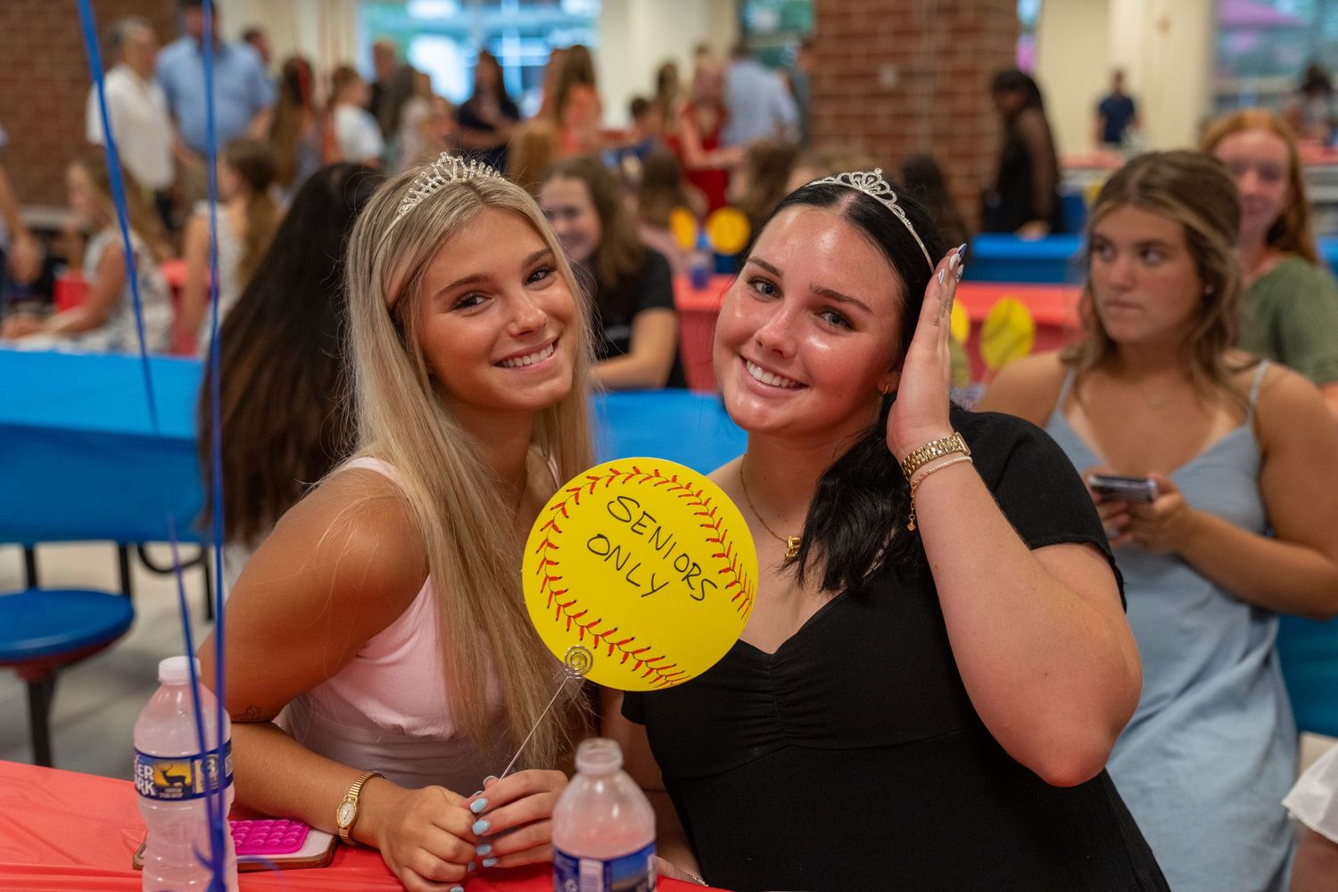 Two smiling young women pose together at a party, one holding a yellow softball sign, wearing tiaras and festive attire.