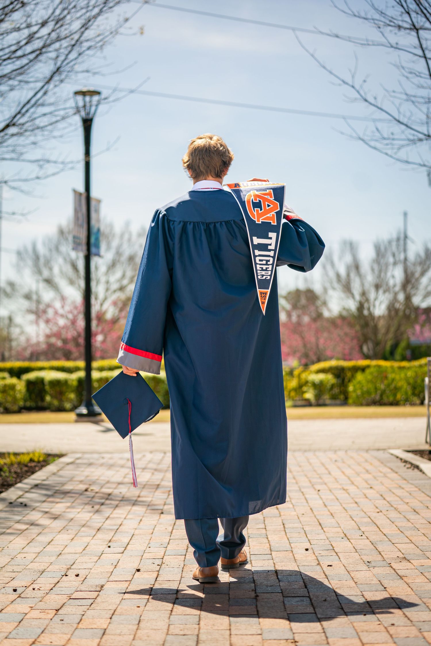A graduate in a navy blue cap and gown walks across a brick courtyard on a spring day with trees in the background.