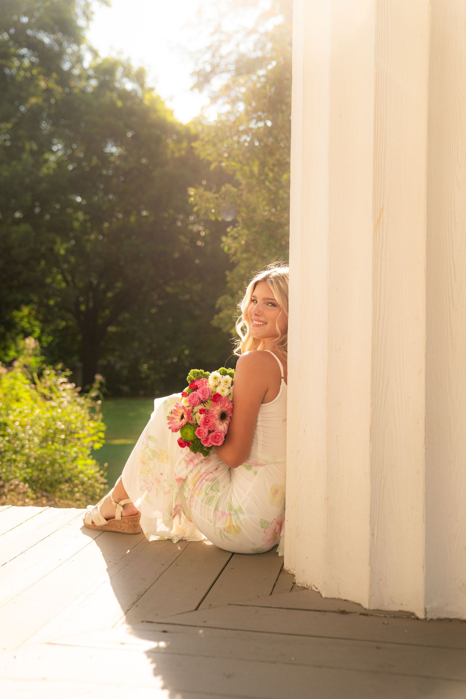 Blonde woman in floral dress sitting by white column holding pink flower bouquet in sunny garden setting.