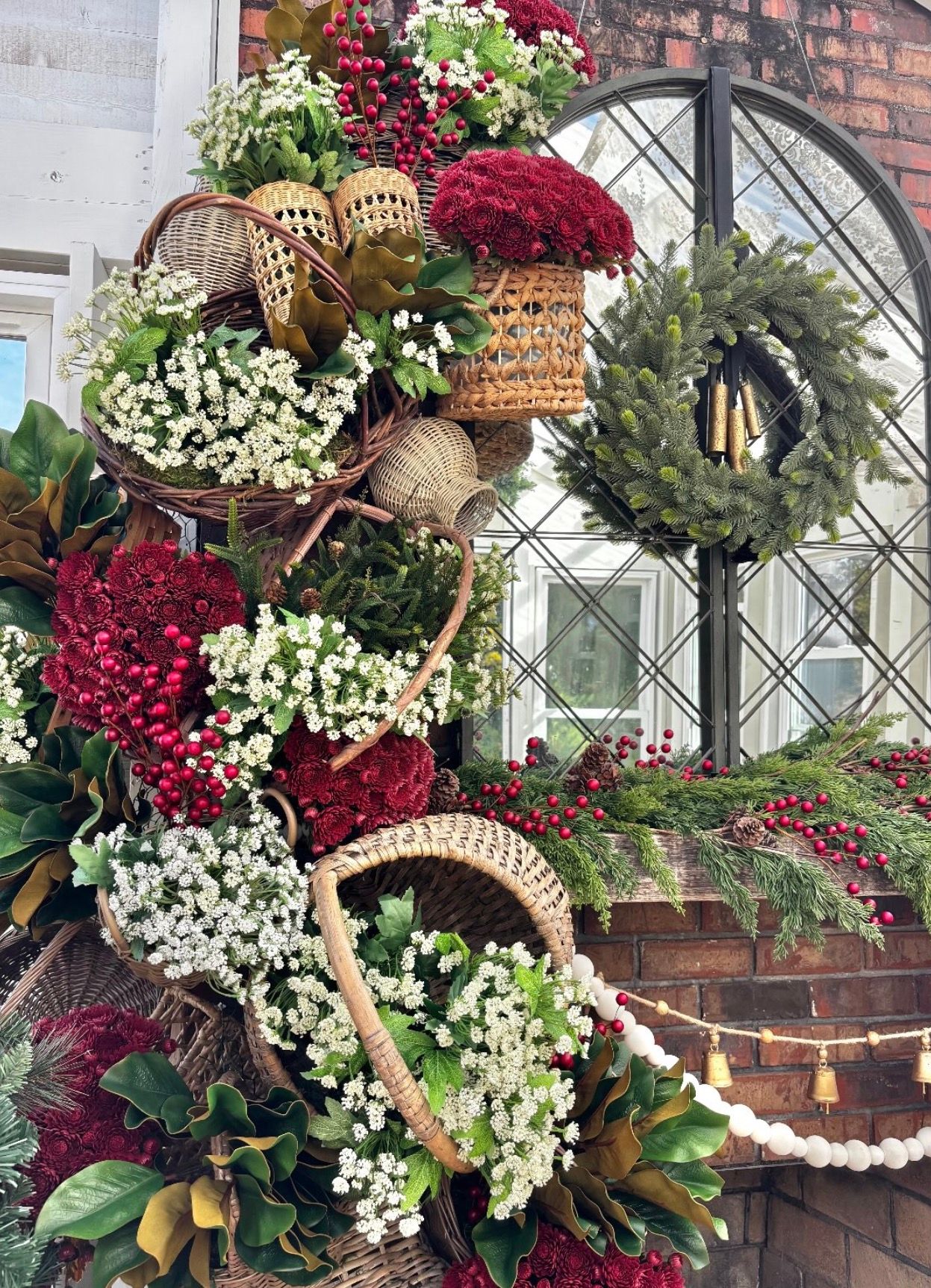 Rustic holiday display with woven baskets of white and red blooms, greenery, a pine wreath, and string lights by an arched window.