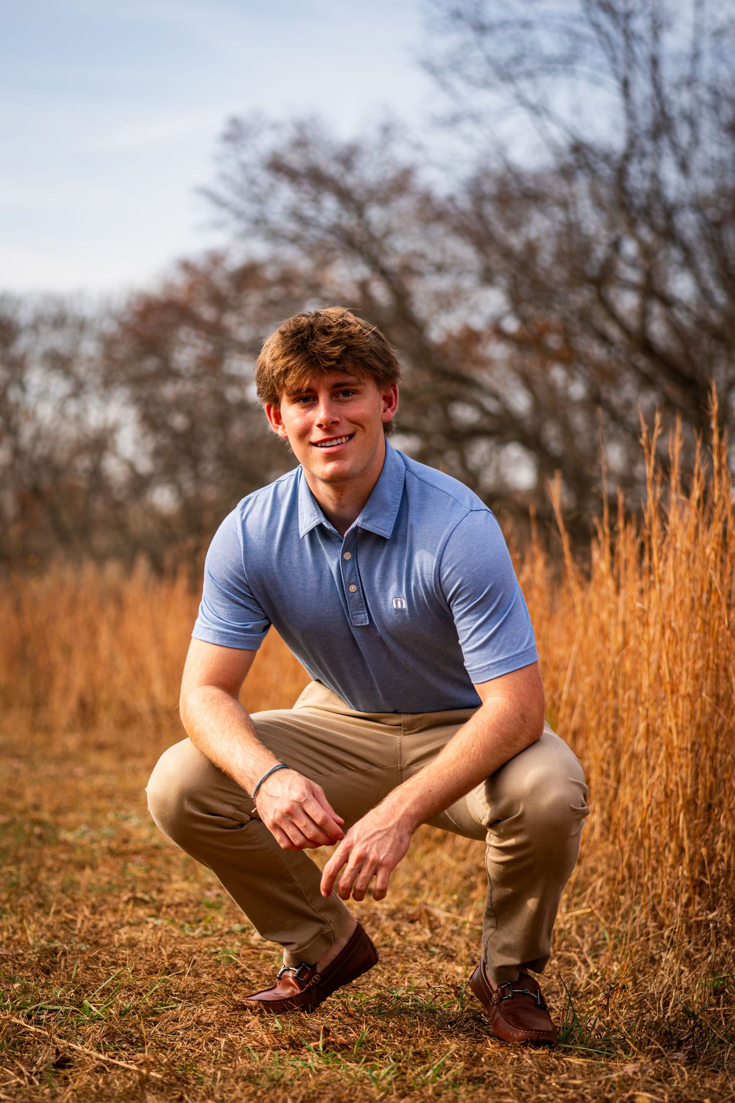A young person in a blue polo shirt crouches in golden autumn grass with trees in the background during sunset.