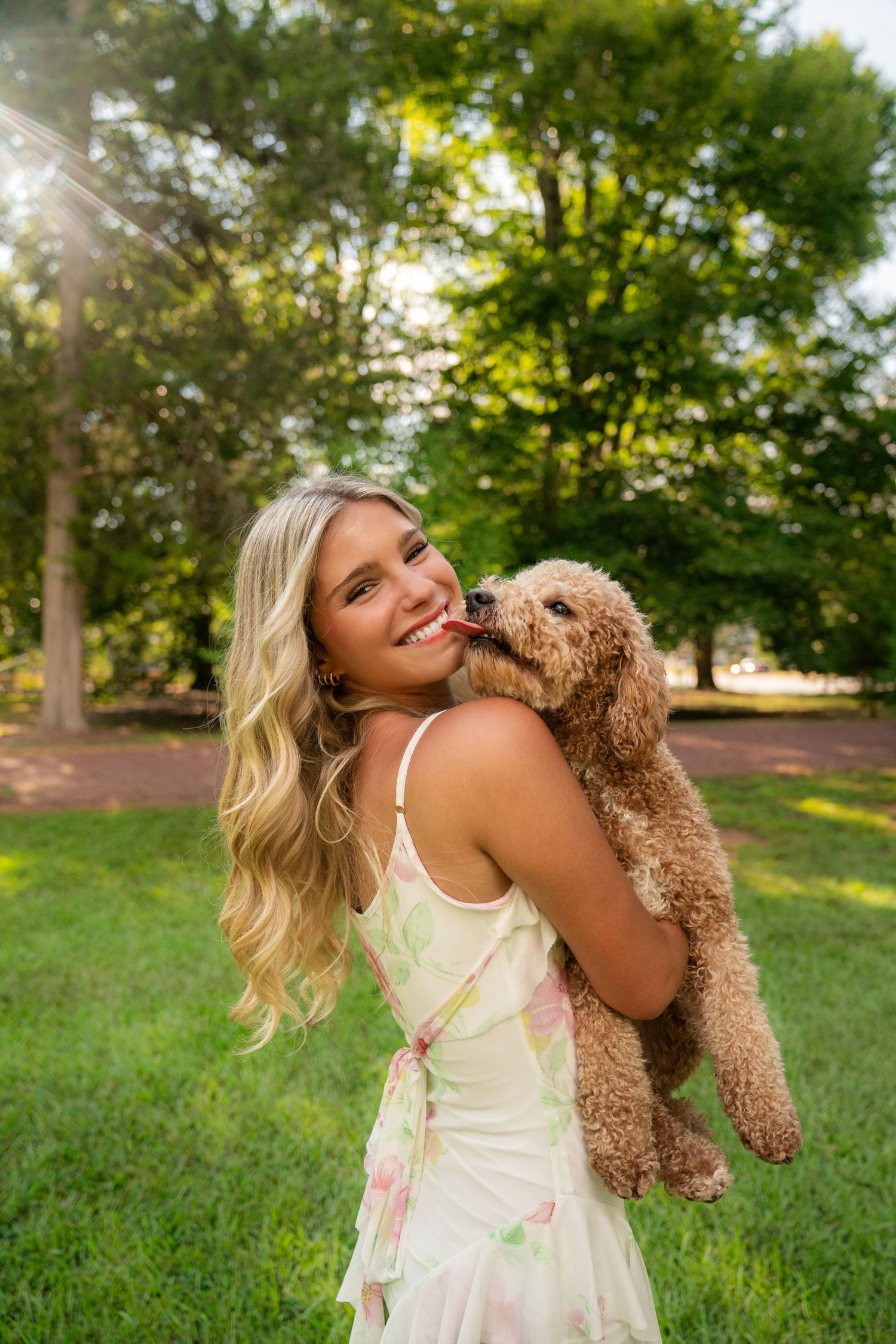 Smiling blonde woman in floral dress holding a golden doodle puppy licking her cheek in a sunny park.