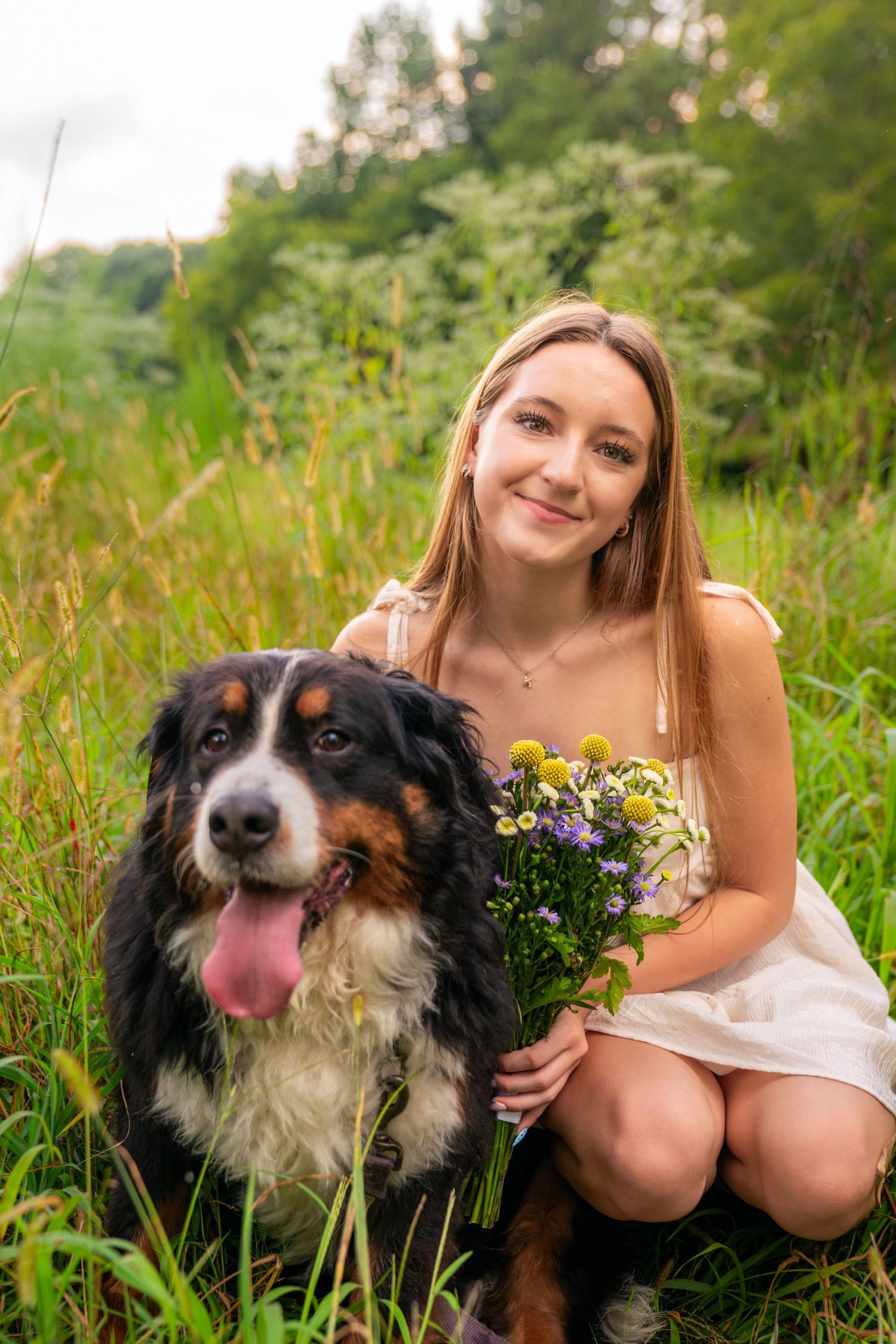Young woman in white dress holds wildflowers while posing with a Bernese Mountain Dog in a lush green field.