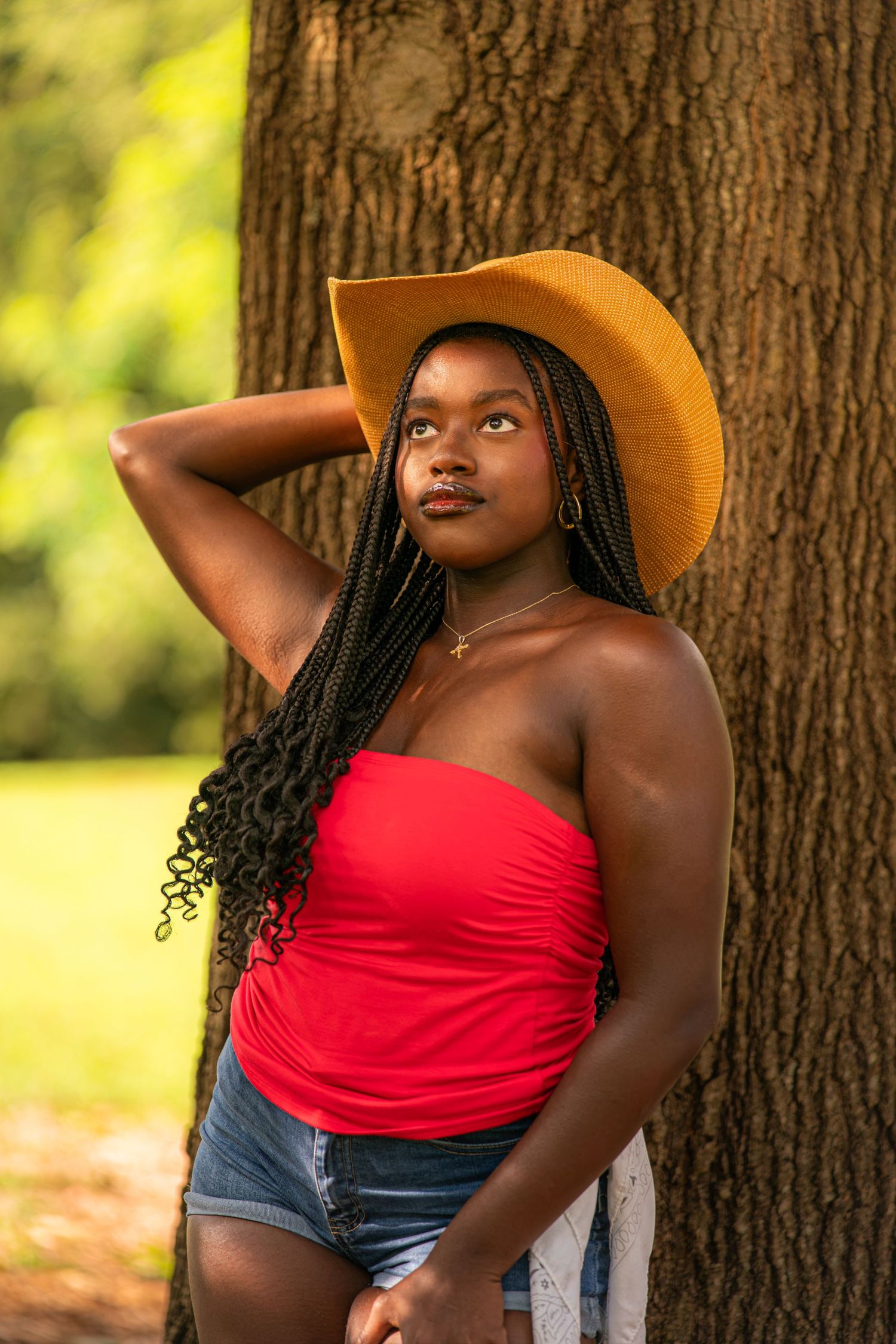 Young Black woman in red tube top, denim shorts, and tan cowboy hat leans against tree in sunny park.