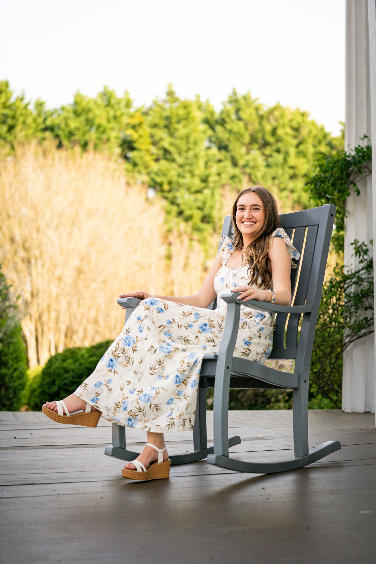 A smiling young woman wearing a floral dress is sitting in a rocking chair on a wooden deck surrounded by greenery.