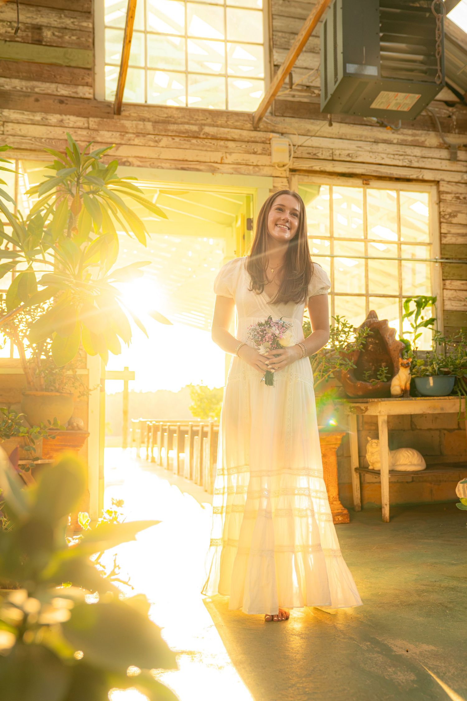 Smiling bride in white gown holding bouquet, bathed in golden sunlight inside a rustic greenhouse.