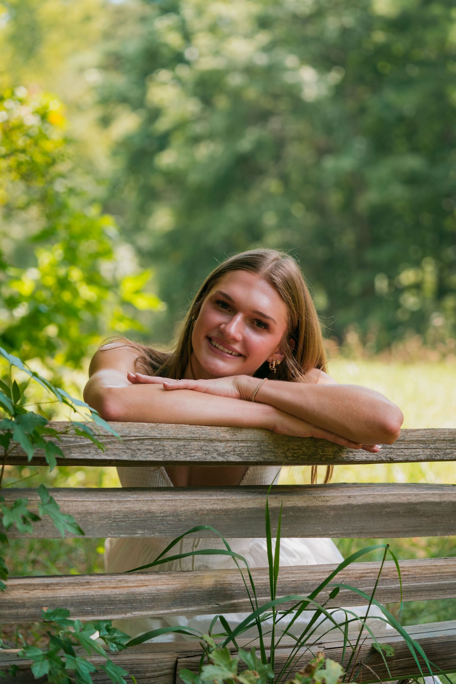 Smiling young woman resting arms on rustic wooden fence surrounded by lush green trees outdoors.