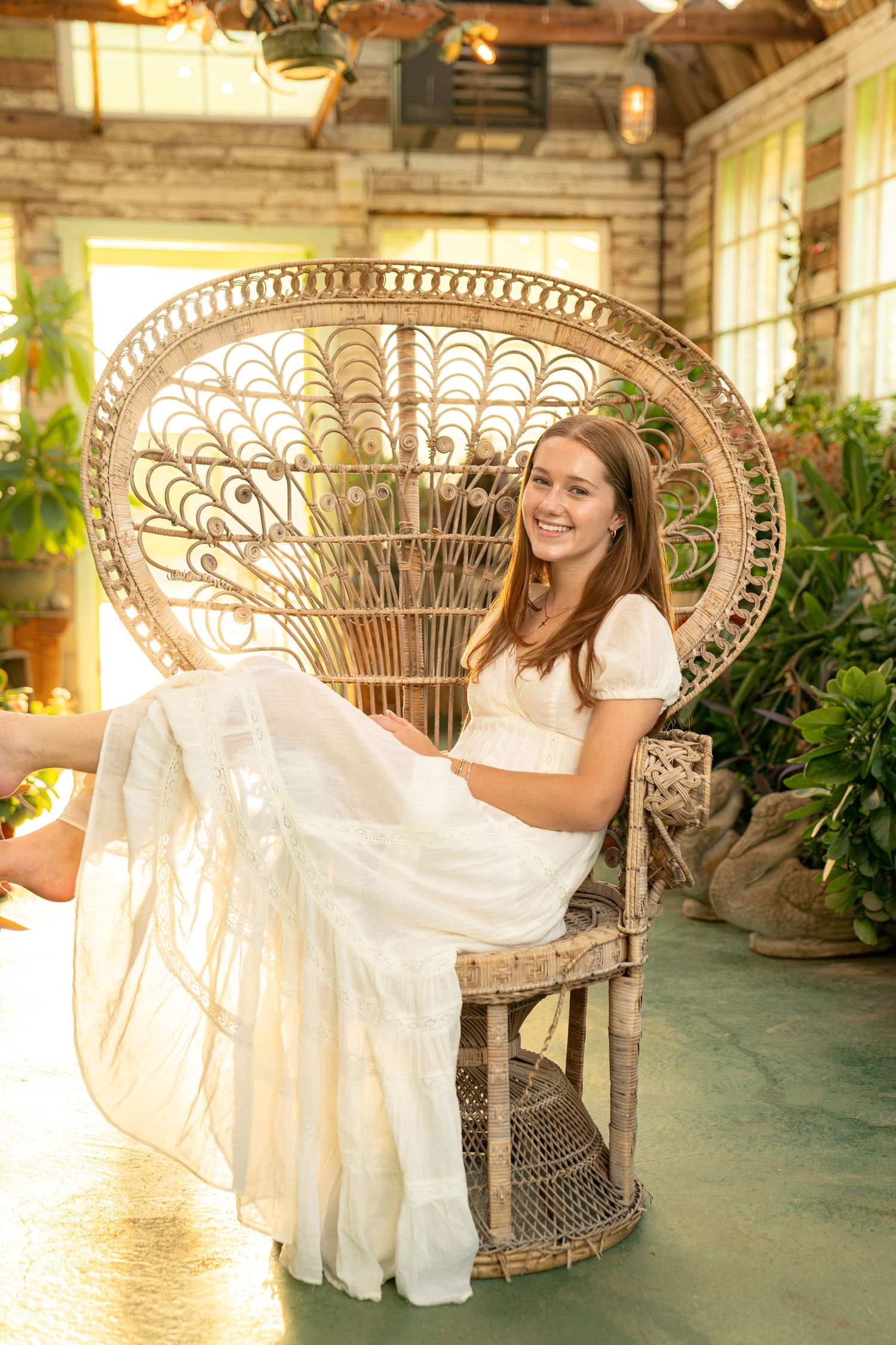 Young woman in white dress sitting in a wicker peacock chair, smiling in a sunlit greenhouse setting.
