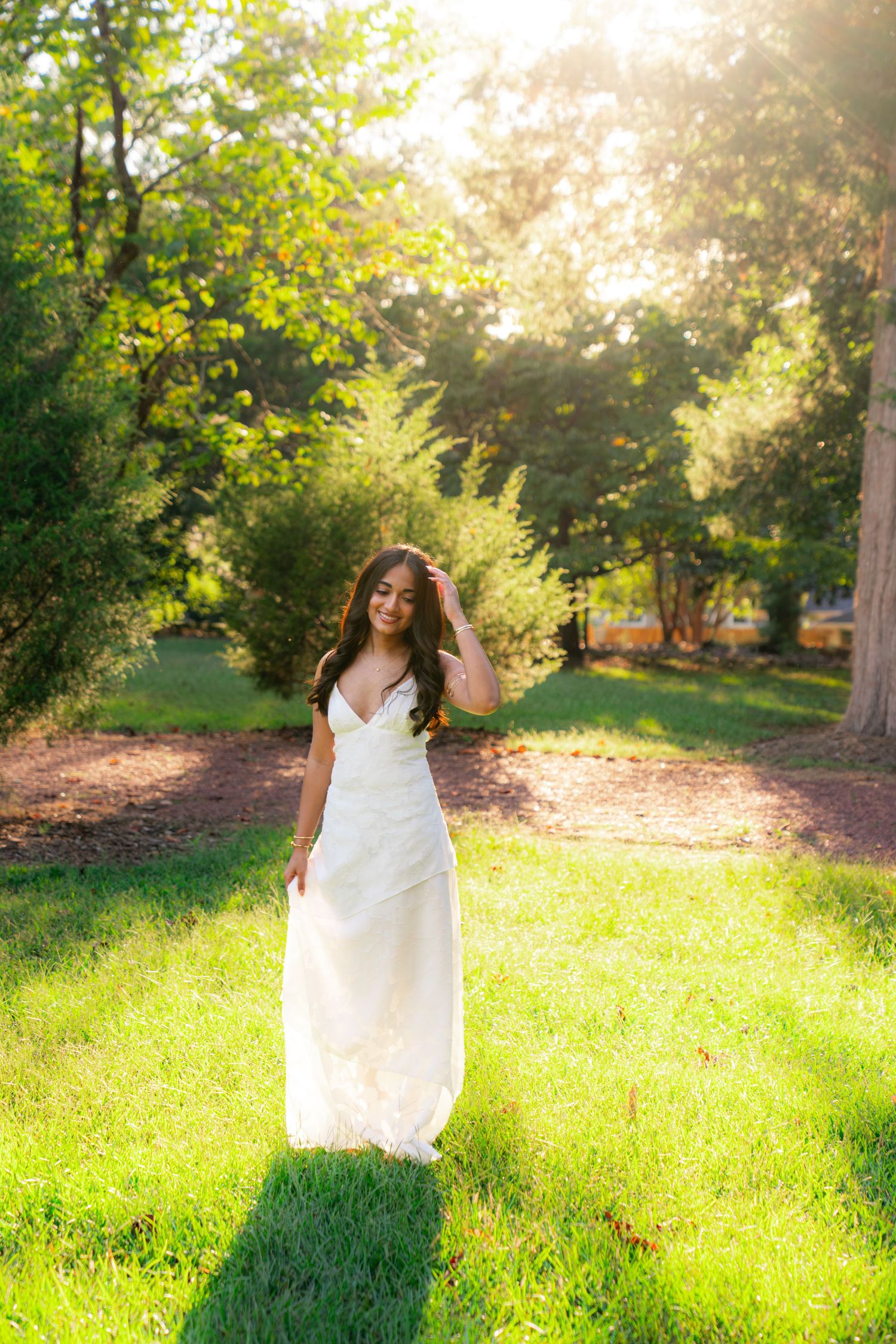 Woman in a flowing white dress walks across a sunlit green lawn, one hand in her long dark hair, surrounded by trees.