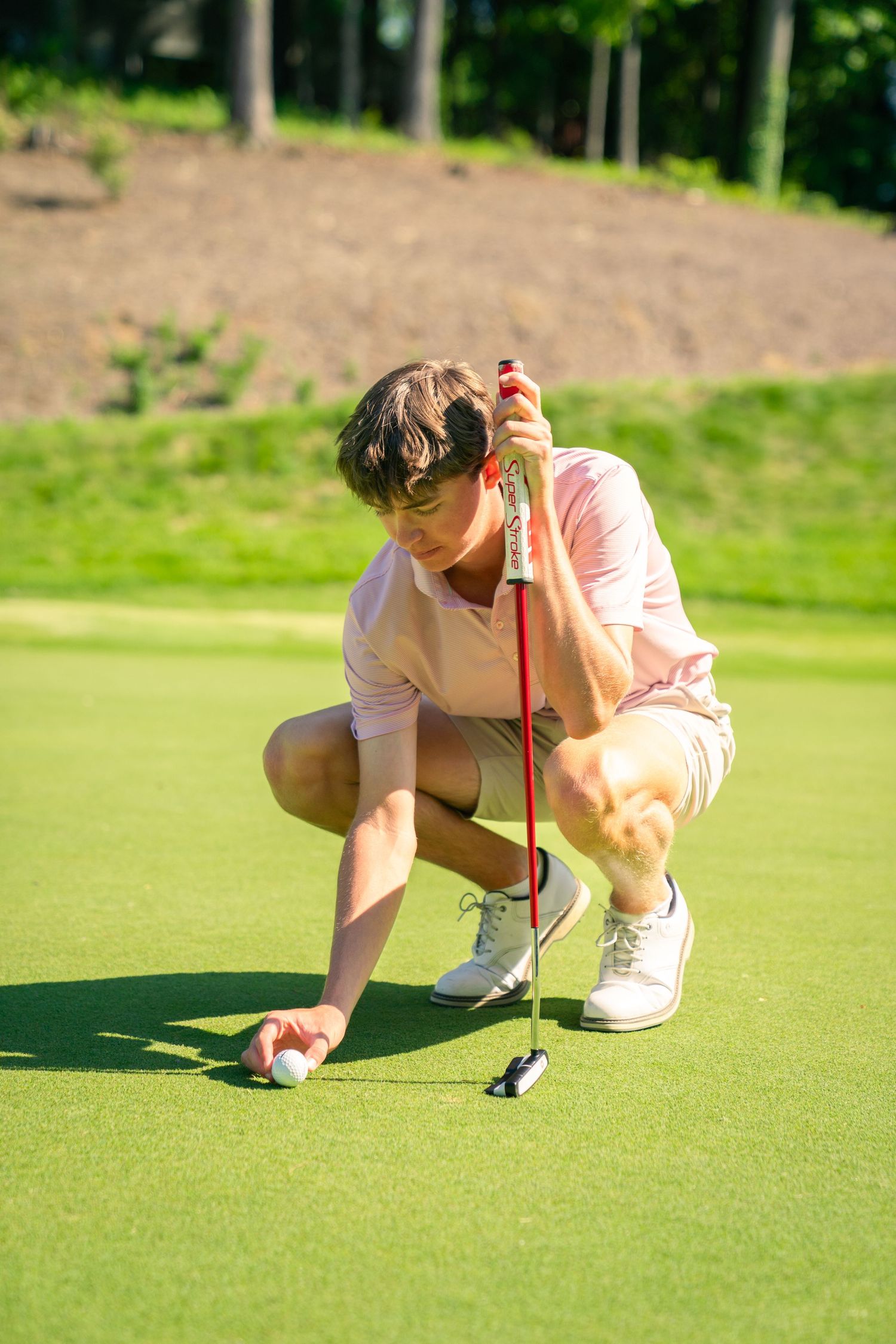 Young golfer in pink shirt crouching on green, carefully placing golf ball while holding red putter on sunny day.