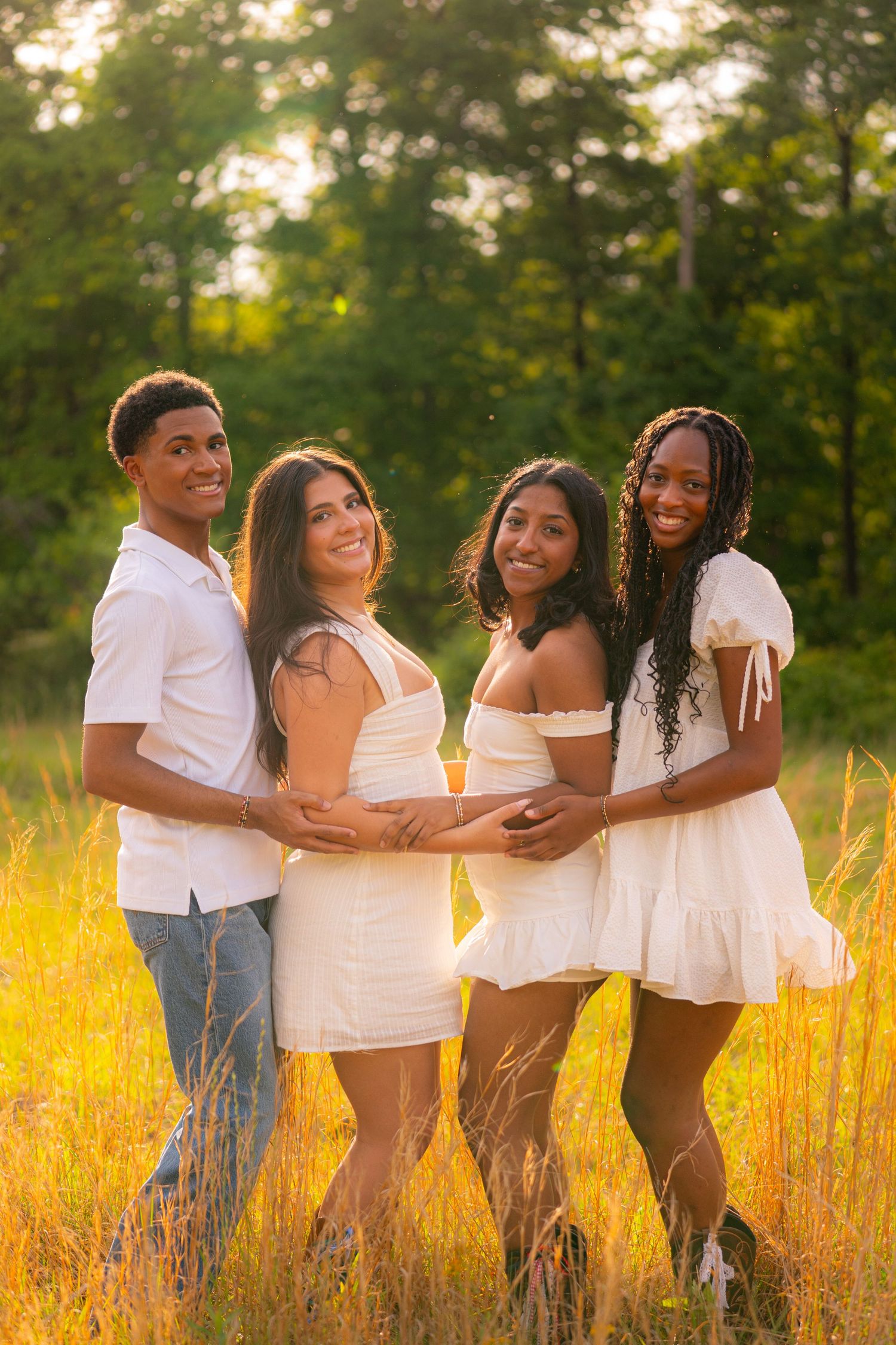 Four young adults in white outfits pose together in a golden sunlit field with lush green trees behind them.