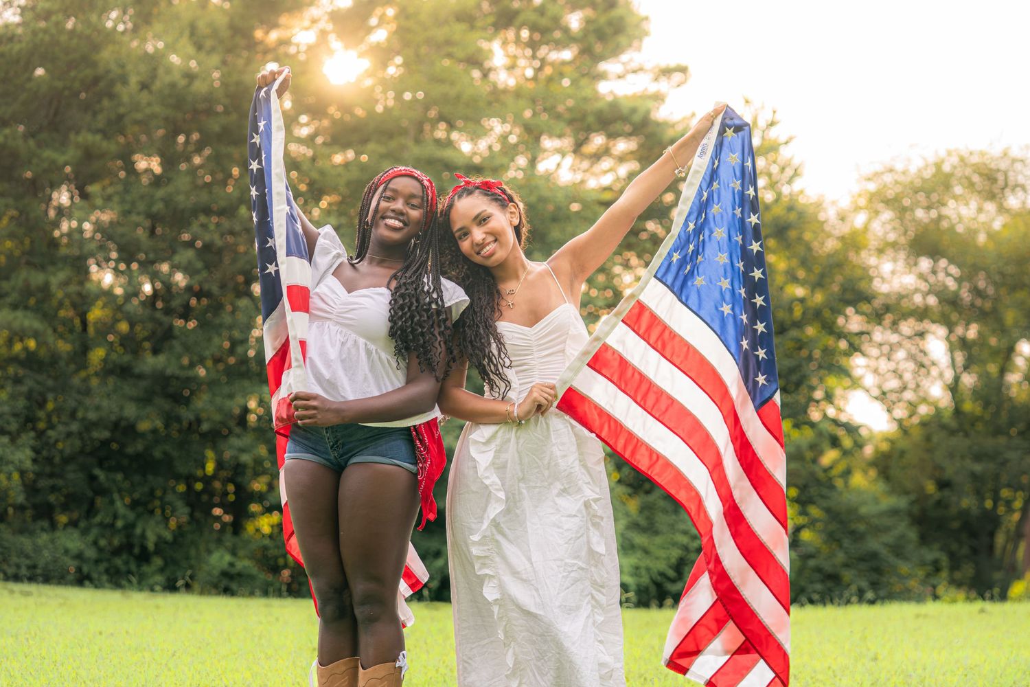 Two women holding an American flag outdoors on green grass, celebrating patriotism with trees in the background.