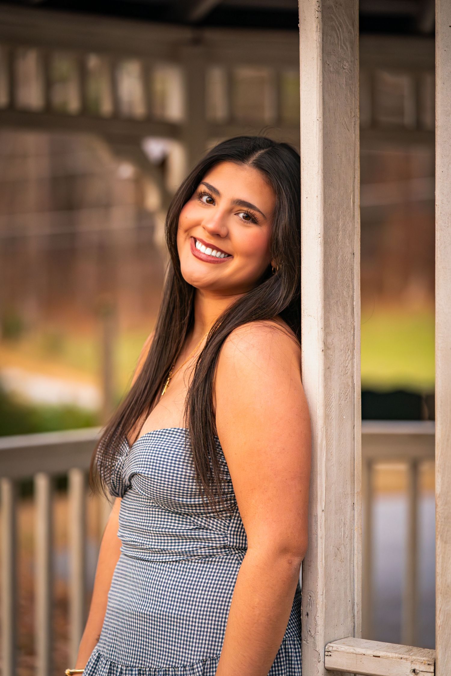 A person in a gingham dress poses against white columns outdoors with a bright smile and long dark hair.