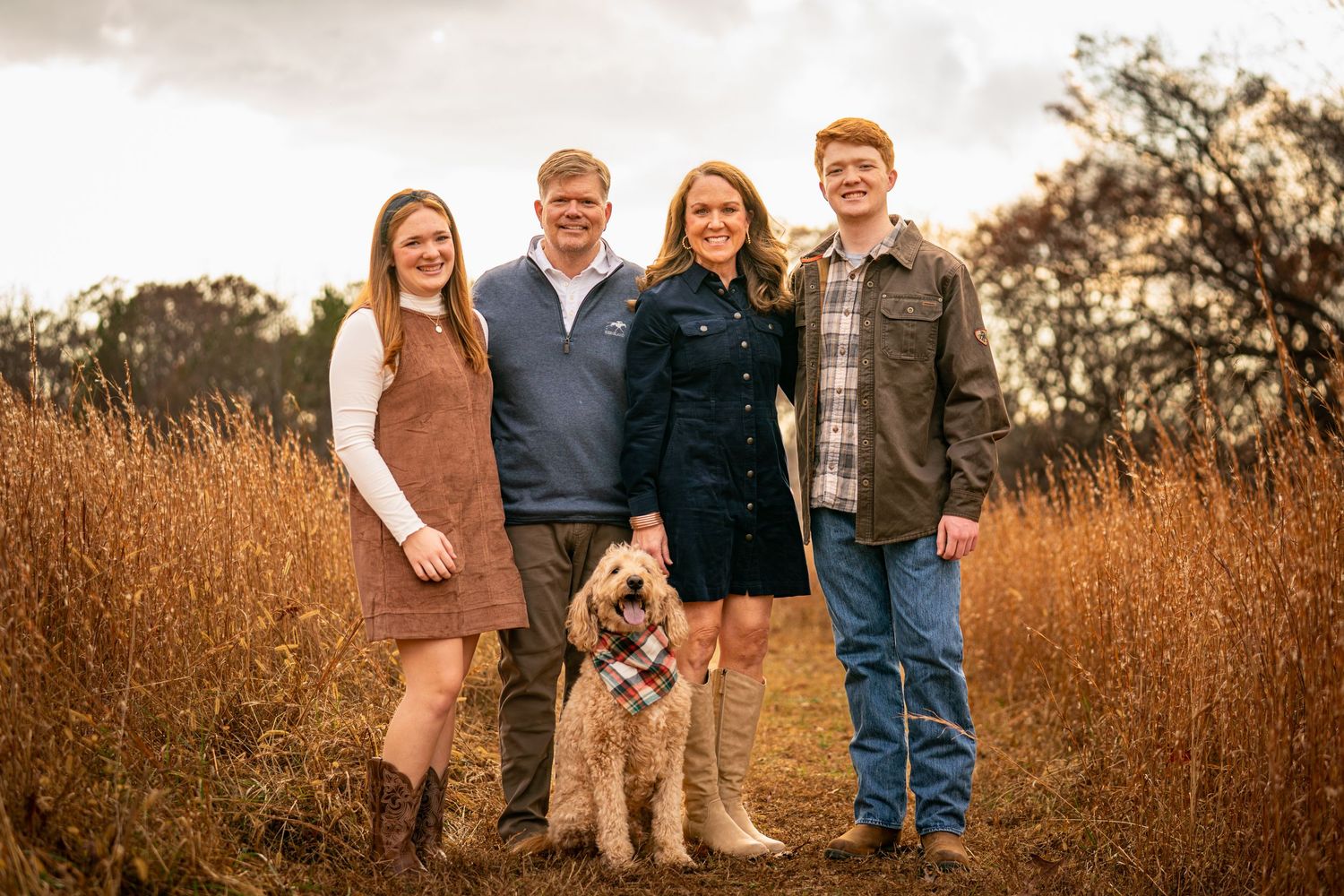 A group stands together with their goldendoodle dog in a golden autumn field at sunset.