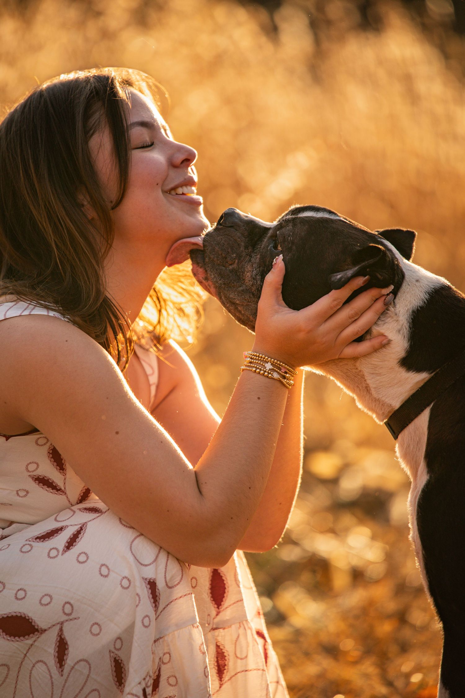 A person in a white dress holds and kisses a black dog against a warm sunset backdrop creating a heartwarming silhouette.