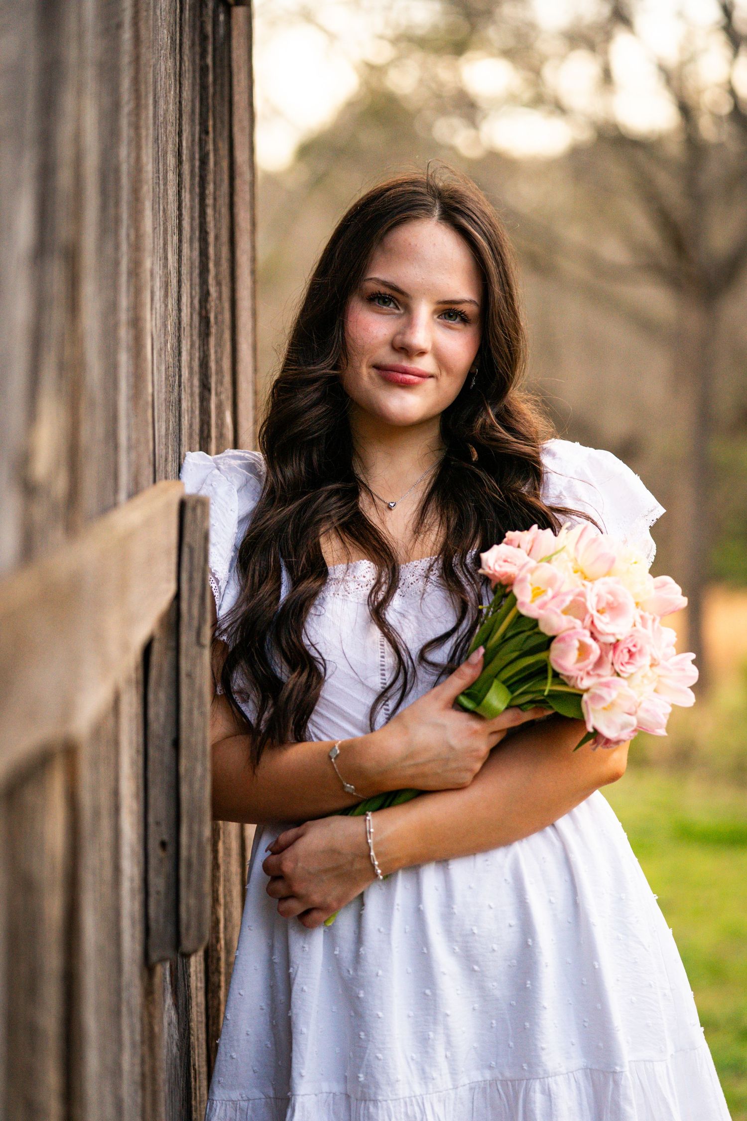 Young woman in white dress holds pink roses against rustic wooden backdrop in outdoor portrait setting.