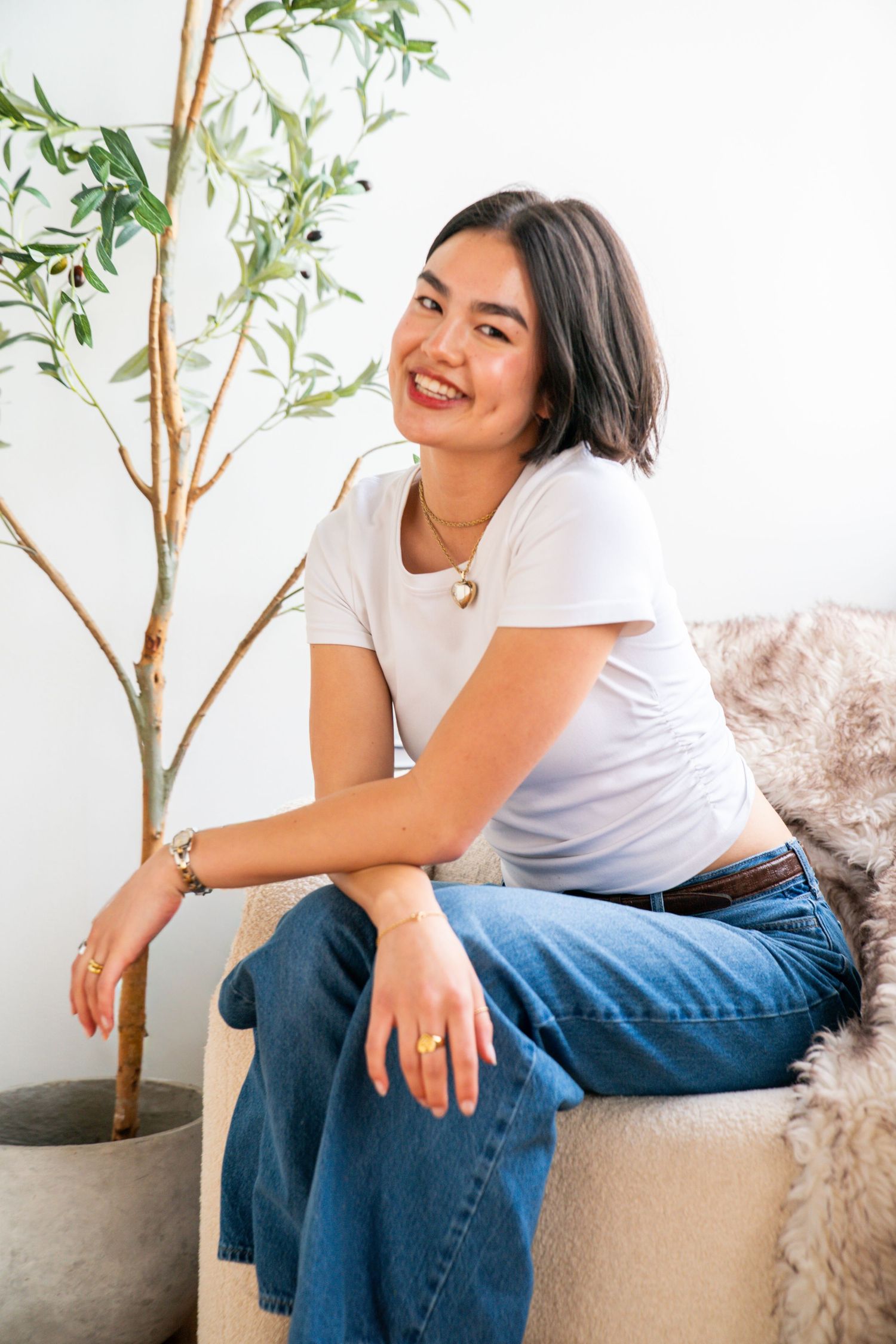 Smiling young woman in white tee and jeans sits on a couch beside a potted olive tree, wearing gold jewelry.