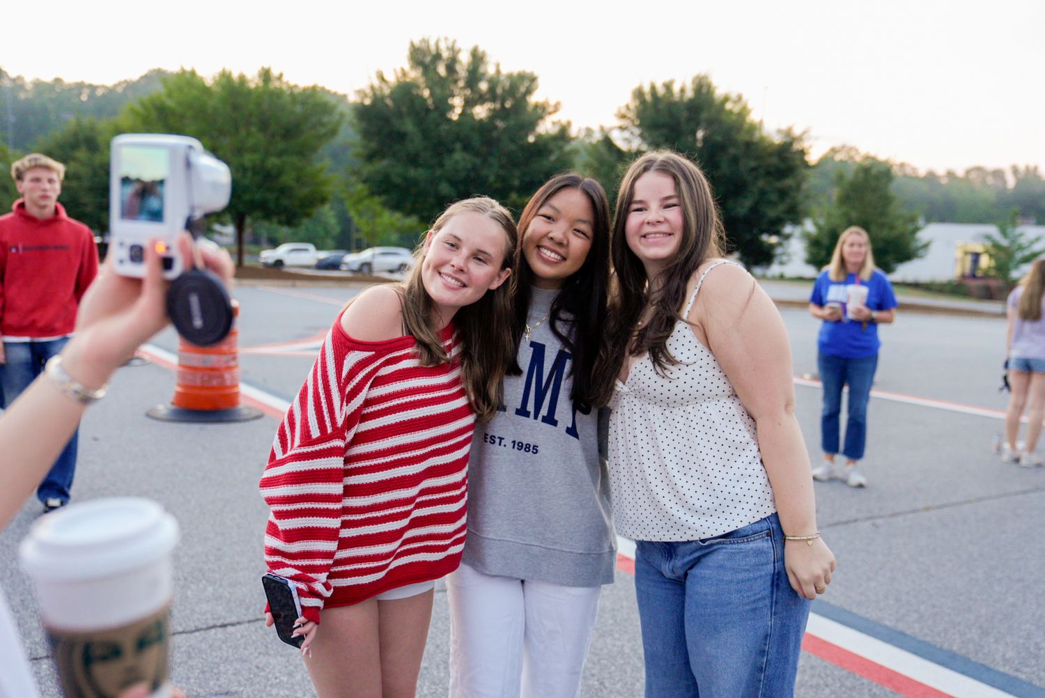 Three smiling teenage girls pose together outdoors at a school event while someone photographs them with a smartphone.