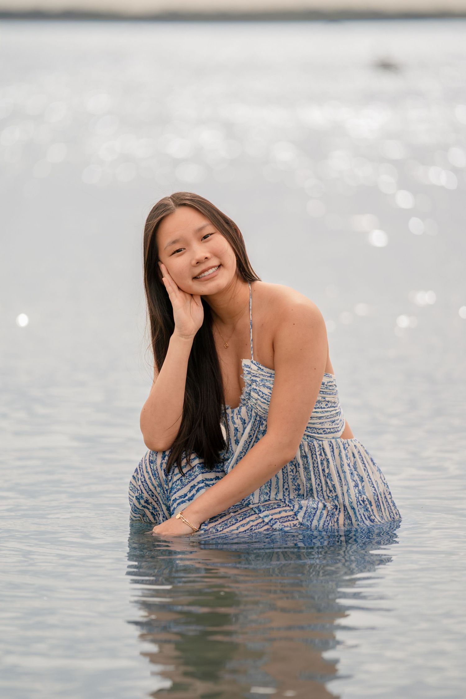 Young Asian woman in blue dress sitting in shallow water, smiling at camera on a sunny beach day.