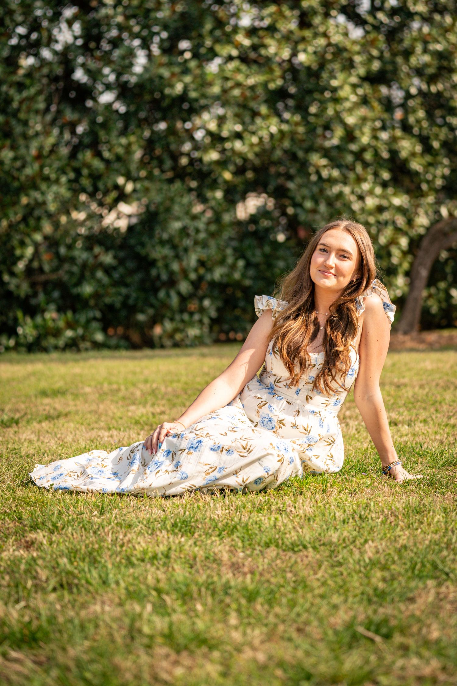 A young woman with long brown hair sits on the grass in a floral dress, smiling at the camera.